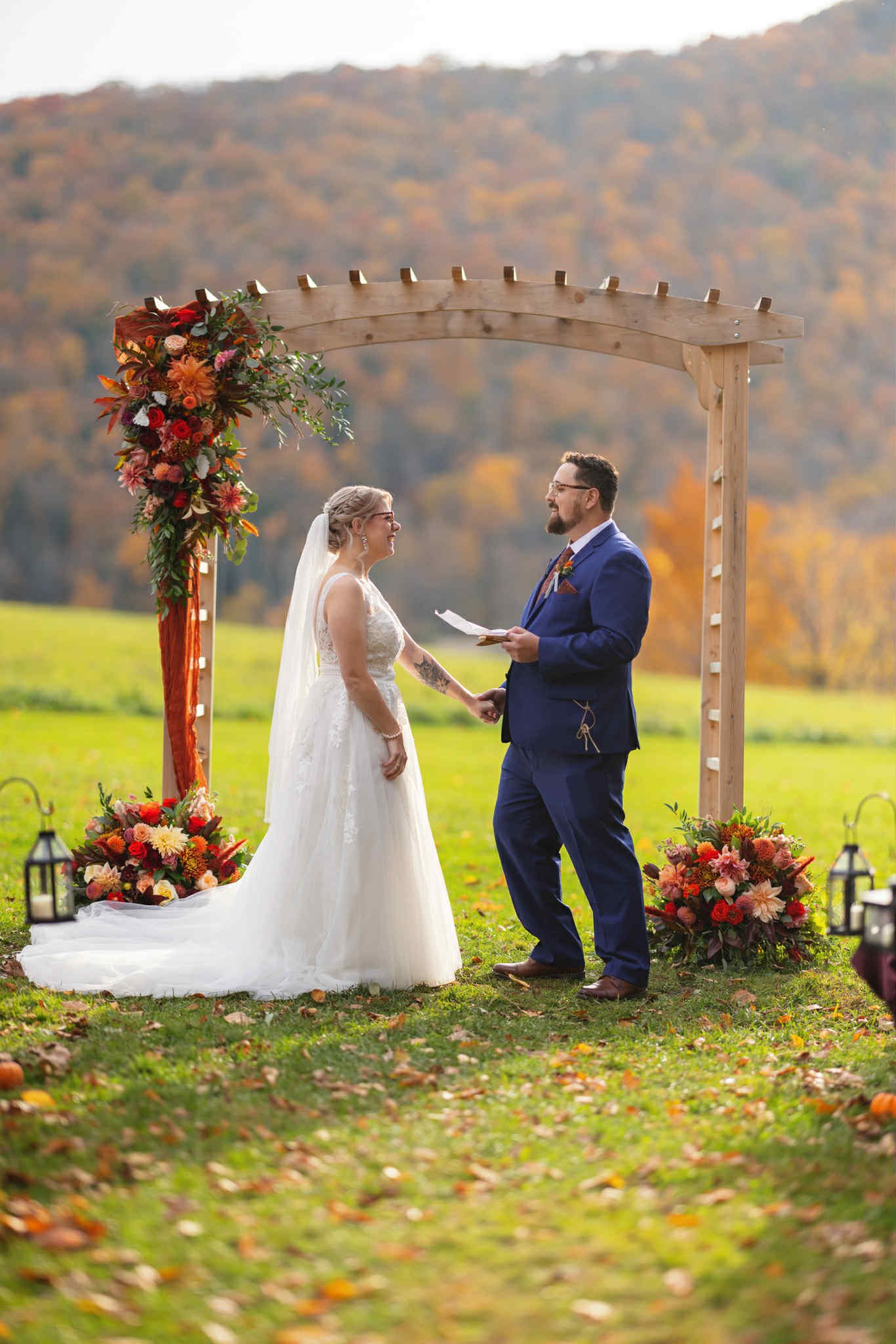 A bride and groom holding hands and facing each other during their outdoor wedding ceremony under a wooden arch decorated with colorful flowers, with two floral arrangements on the ground and lanterns, set against a scenic autumnal landscape.