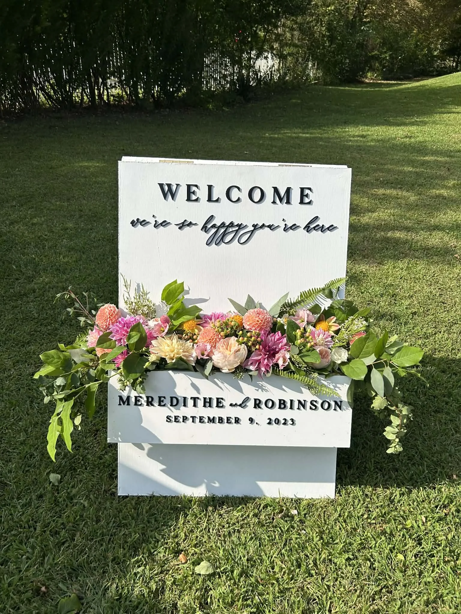 A white welcome sign with black lettering, decorated with pink, peach, and yellow flowers designed by Lot 32 Flower Farm, placed on a grassy area.