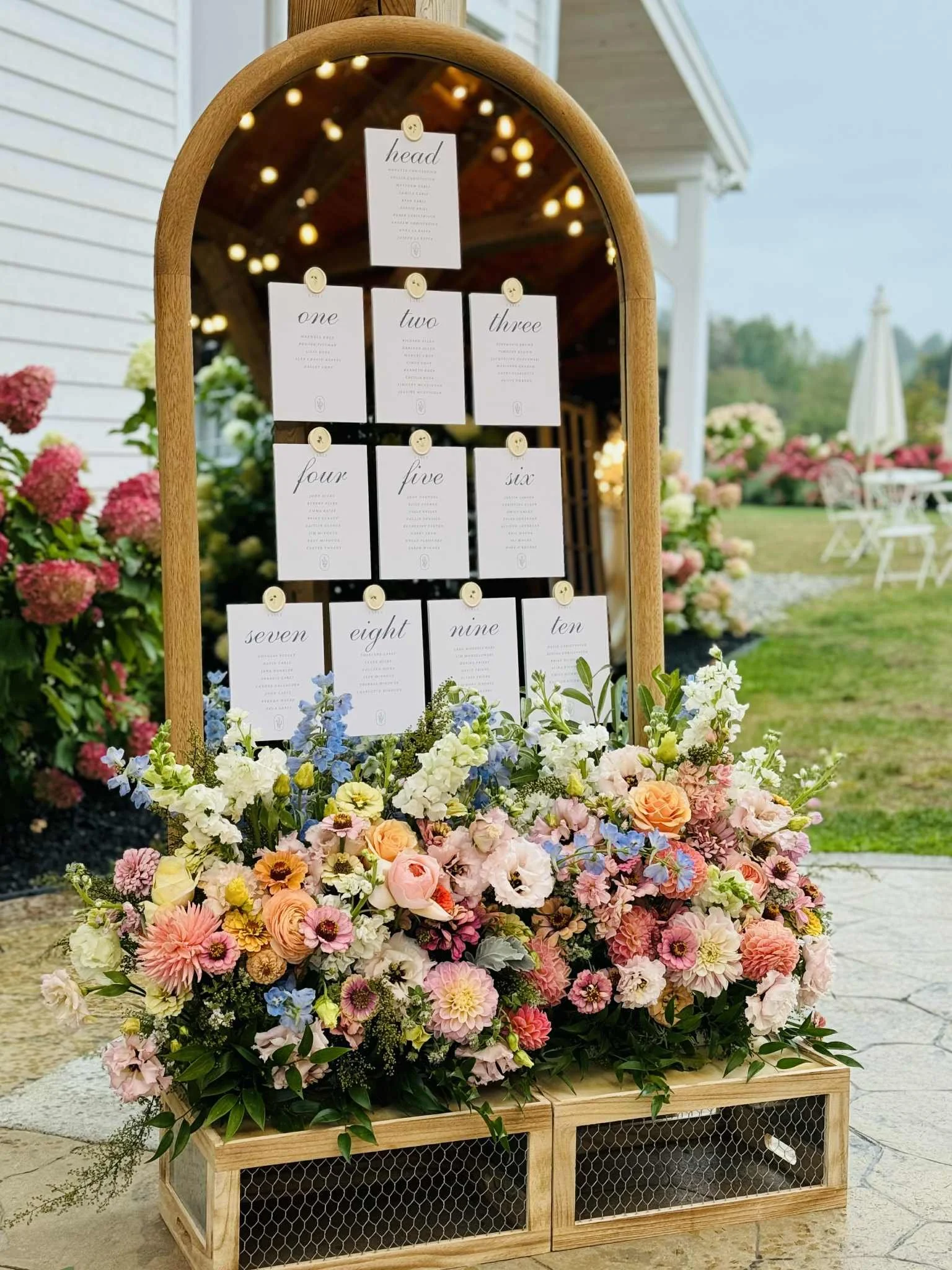 A decorative welcome sign with table numbers at a wedding or event, surrounded by a display of colorful flowers including pink, white, and blue blooms, with an outdoor setting visible in the background.