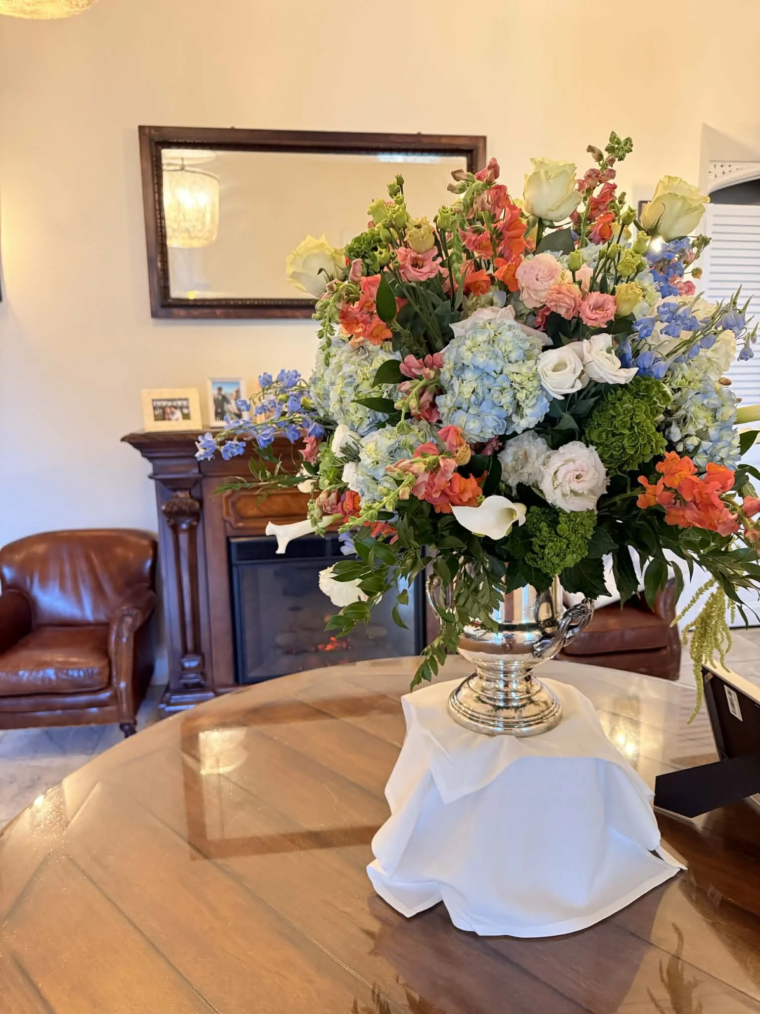 Large floral arrangement with white, pink, orange, blue, and green flowers in a silver vase on a wooden table with a white cloth underneath in a living room.