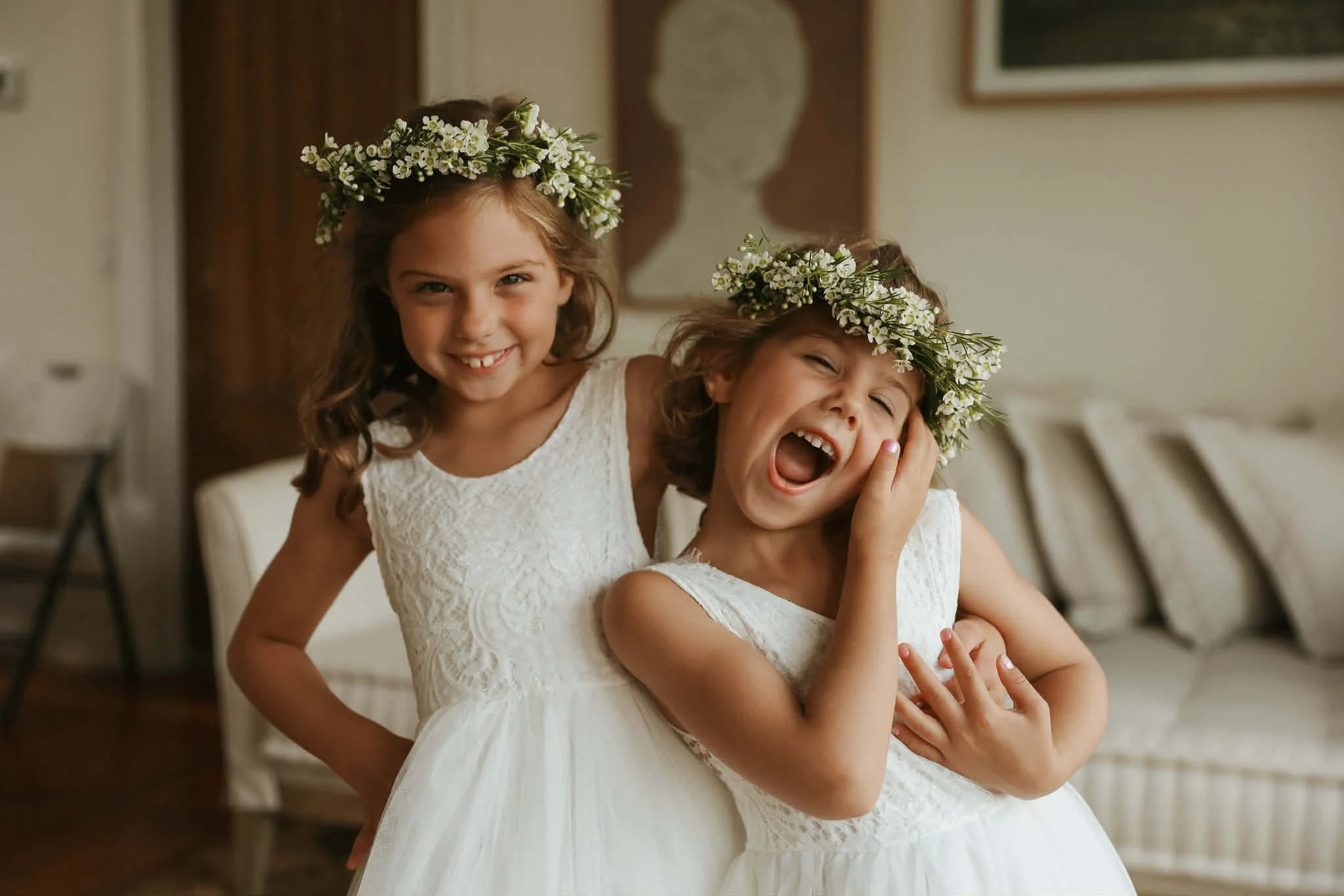 Two young girls wearing white dresses and floral crowns, smiling and laughing indoors.