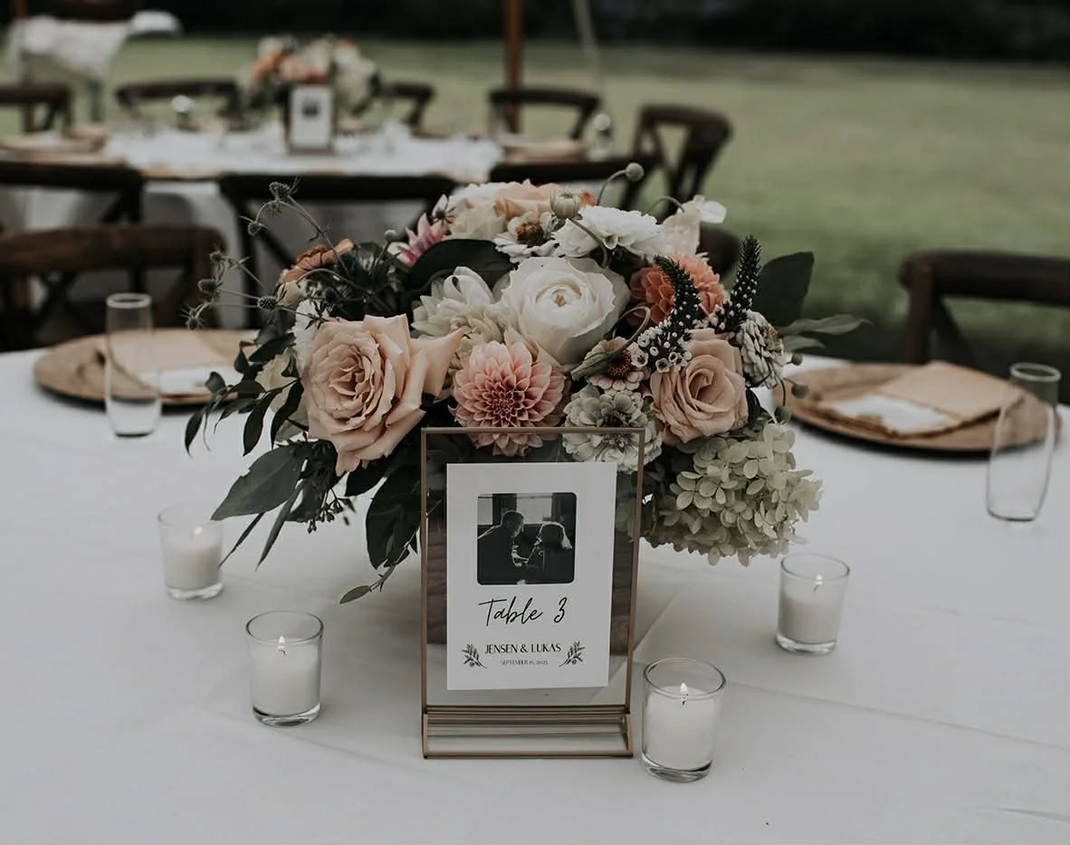 A floral centerpiece with pink, white, and peach flowers on a white table at an outdoor wedding reception, with table number and candles surrounding it.