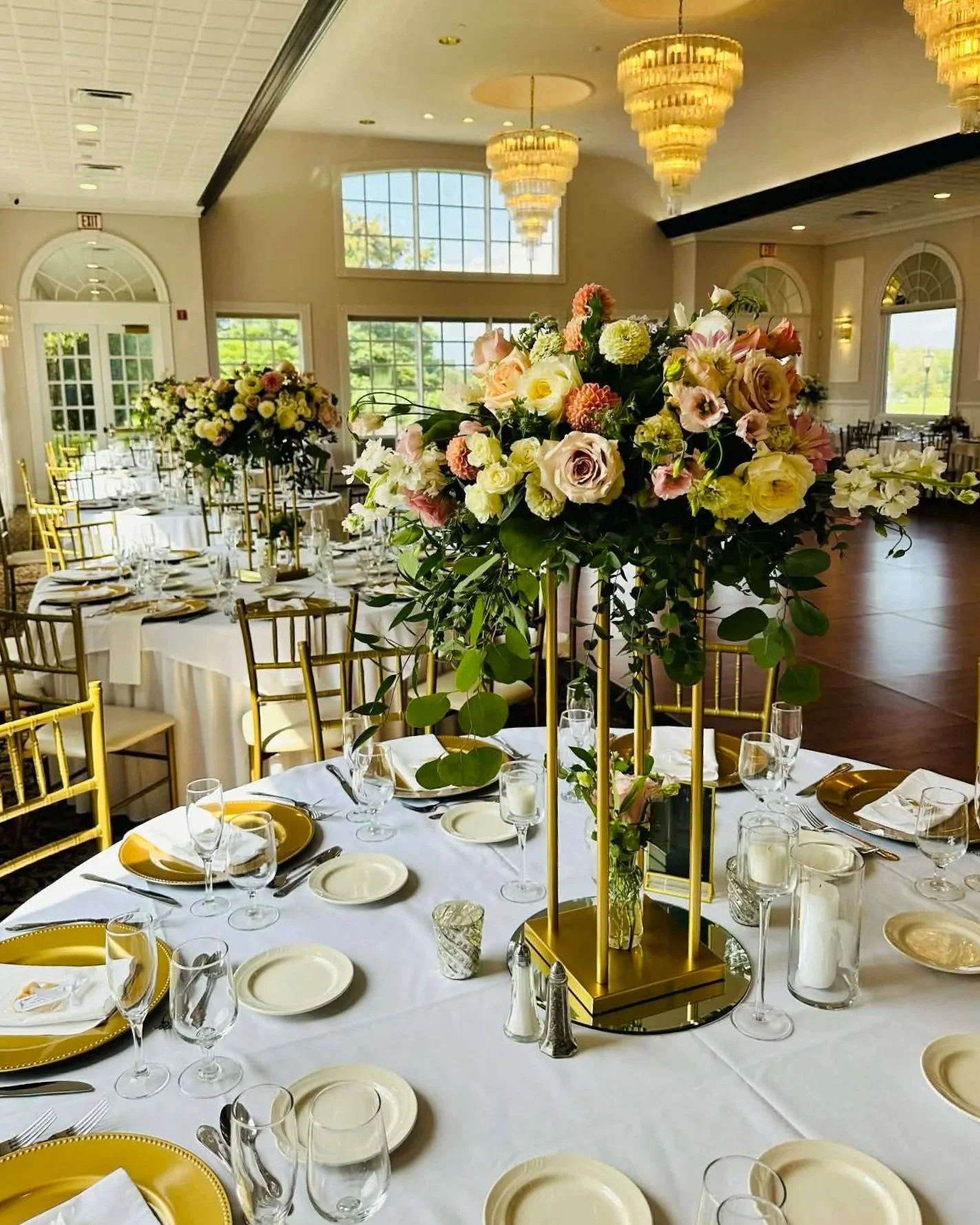 Pastel wedding arrangements elevated above round tables on gold pedestals in a ballroom.