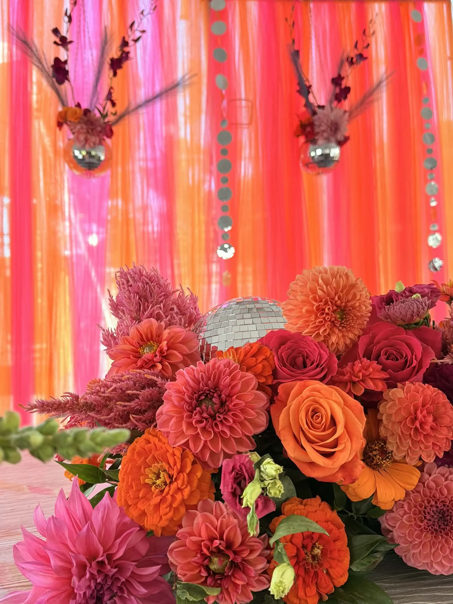 A colorful floral arrangement with pink, orange, and purple flowers on a table in front of a vibrant pink and orange backdrop with hanging silver disco balls.