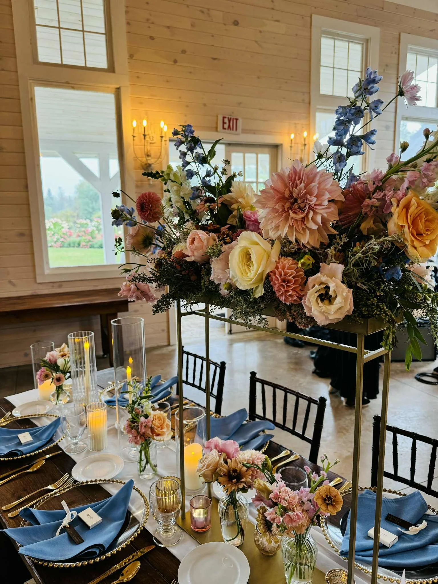 Elegant table setting with floral arrangements, candles, and blue napkins in a rustic wooden room with large windows and warm lighting.
