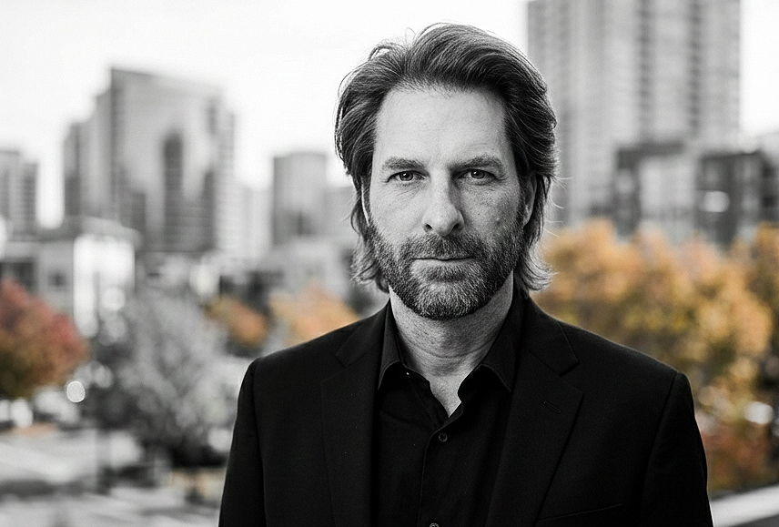 Black and white photo of a man with a beard and medium-length hair, wearing a dark suit and shirt, standing outdoors in an urban area with buildings and trees with fall foliage in the background.