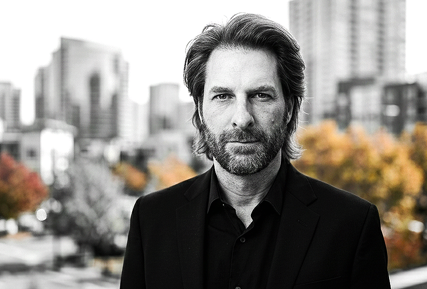 A man with shoulder-length hair and a beard wearing a black suit and shirt, standing outdoors in an urban setting with blurred buildings and trees with autumn foliage in the background.