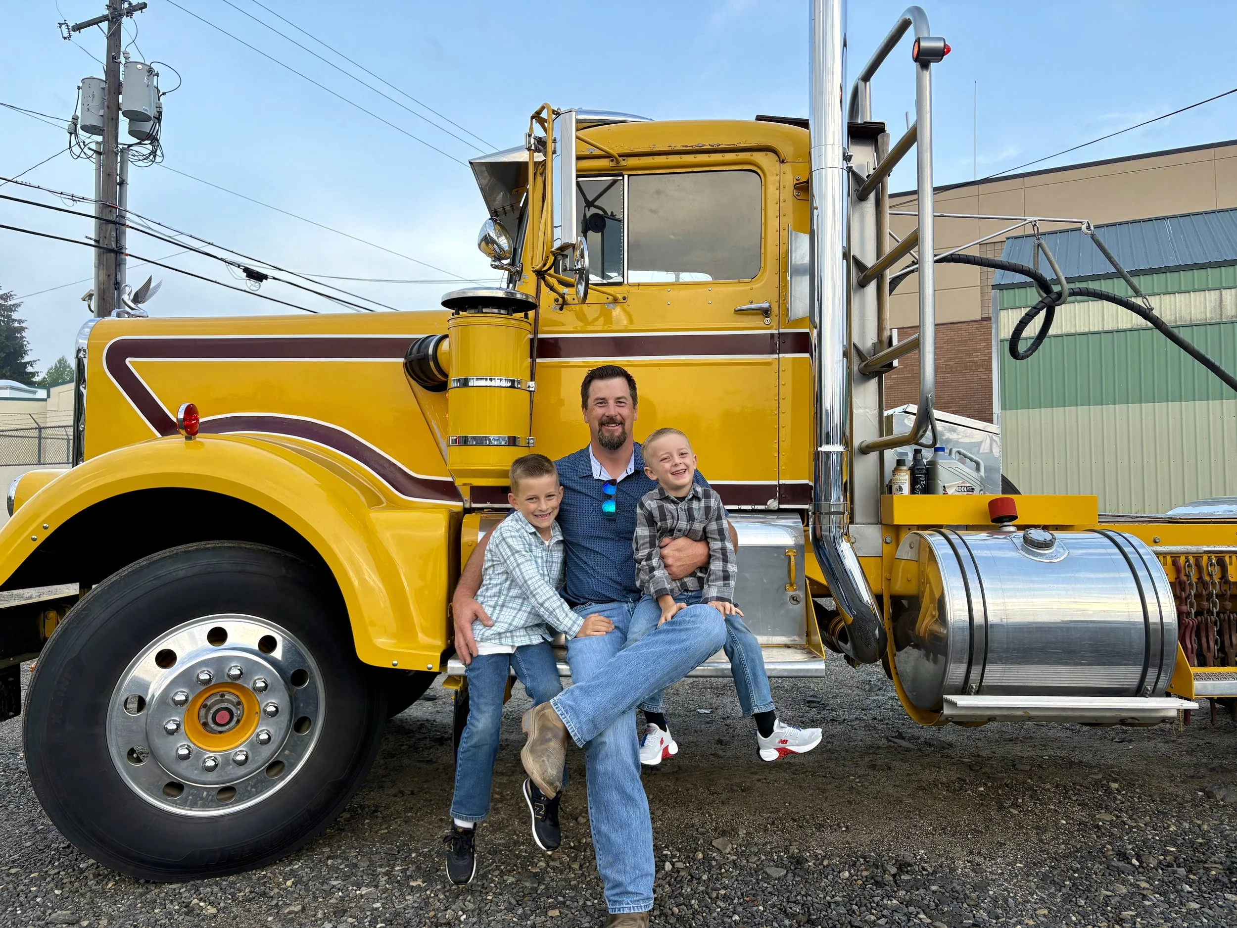 A man with two children sitting on his lap in front of a yellow semi-truck, smiling and posing for the camera.
