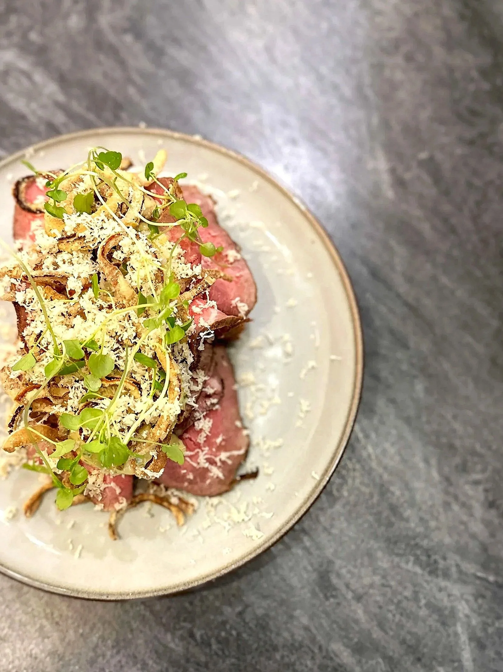 Thinly sliced cooked beef topped with shredded cheese, fried onions, and microgreens on a ceramic plate.