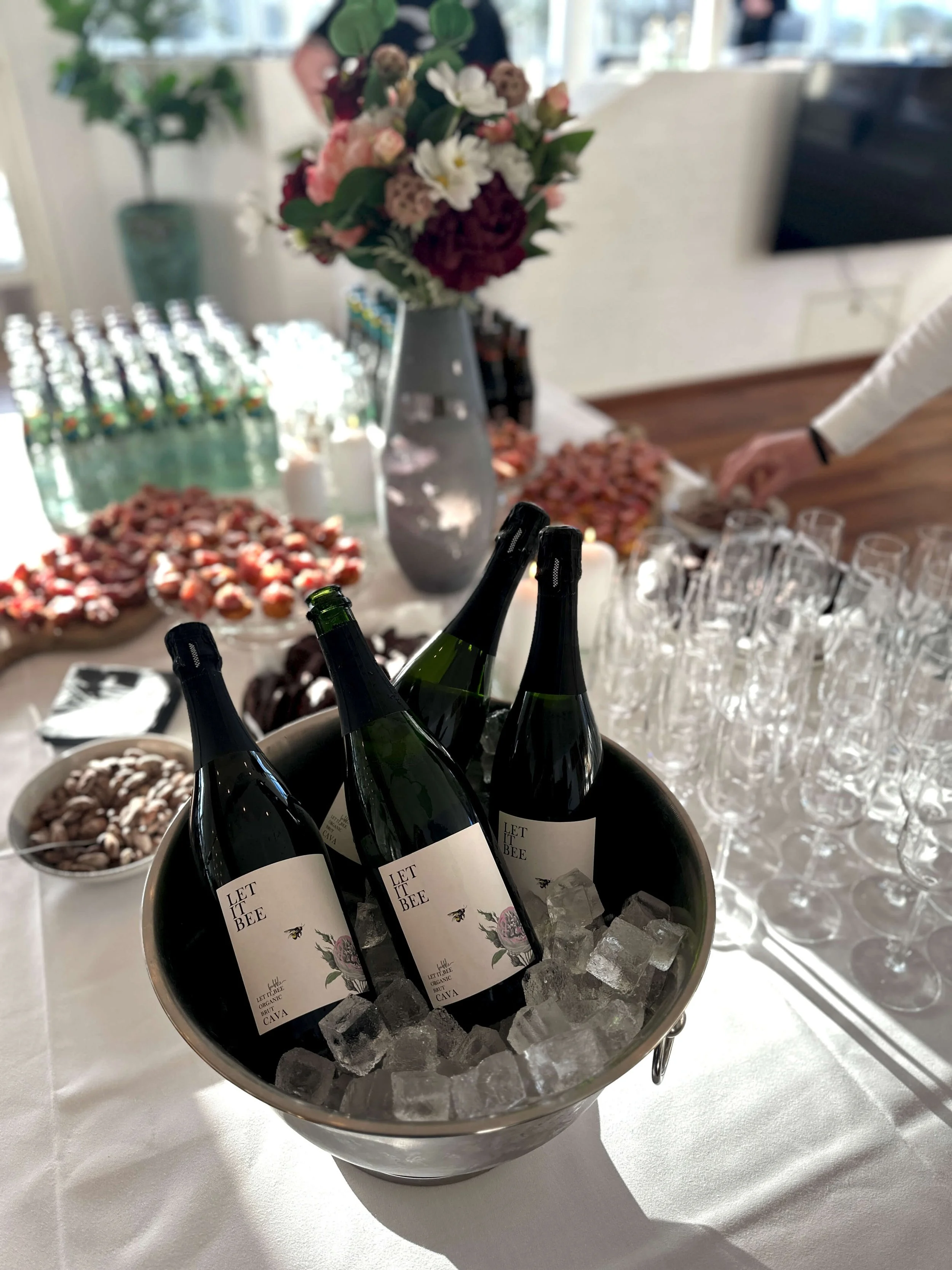 Four bottles of sparkling wine labeled 'Let It Bee' chilling in ice in a silver bucket on a table with various desserts and a flower arrangement in the background.