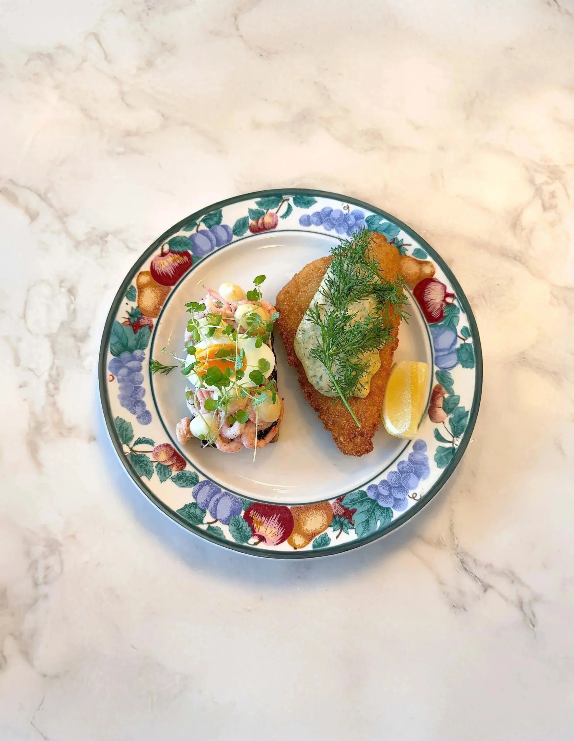 Plate with breaded fish fillet topped with dill and a lemon wedge, served with a gourmet salad and fresh herbs.