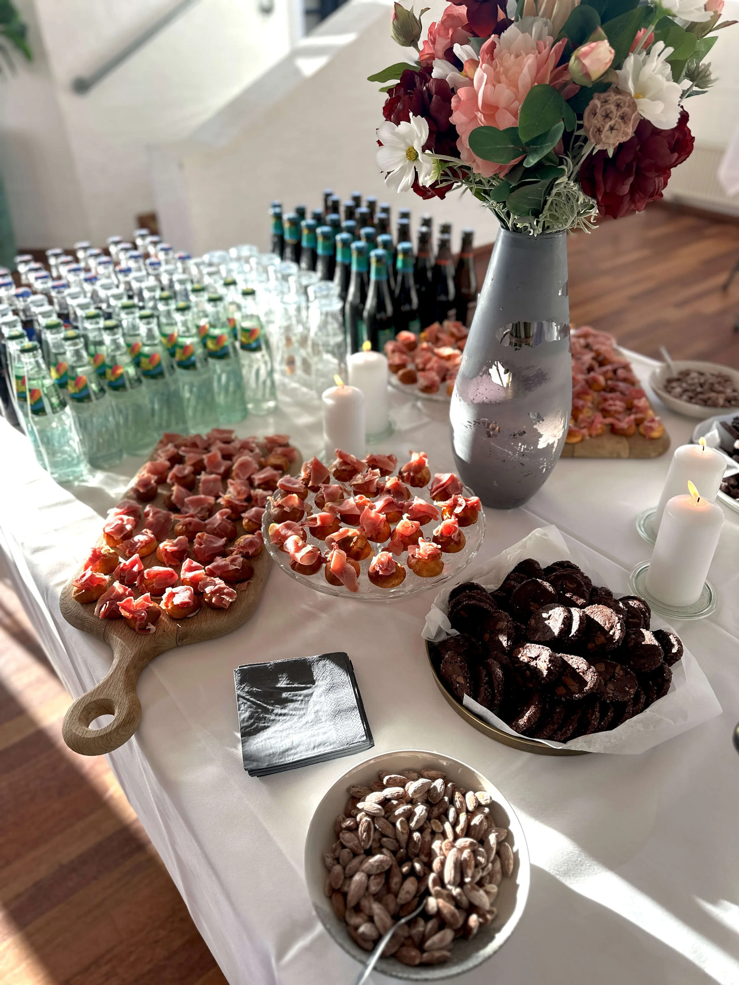 A table set with a large floral bouquet in a gray vase, assorted cookies, nuts, candies, bottles, and candles, ready for a celebration or gathering.