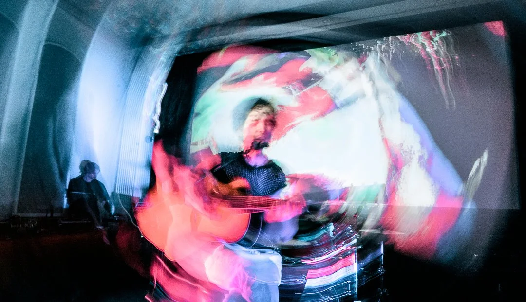 Musician playing guitar and singing on stage with colorful, blurred lights and visuals in the background.