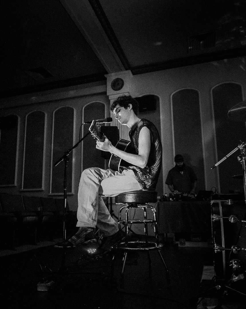 A musician sitting on a stool playing an acoustic guitar and singing into a microphone during a performance, with a DJ or sound engineer in the background.