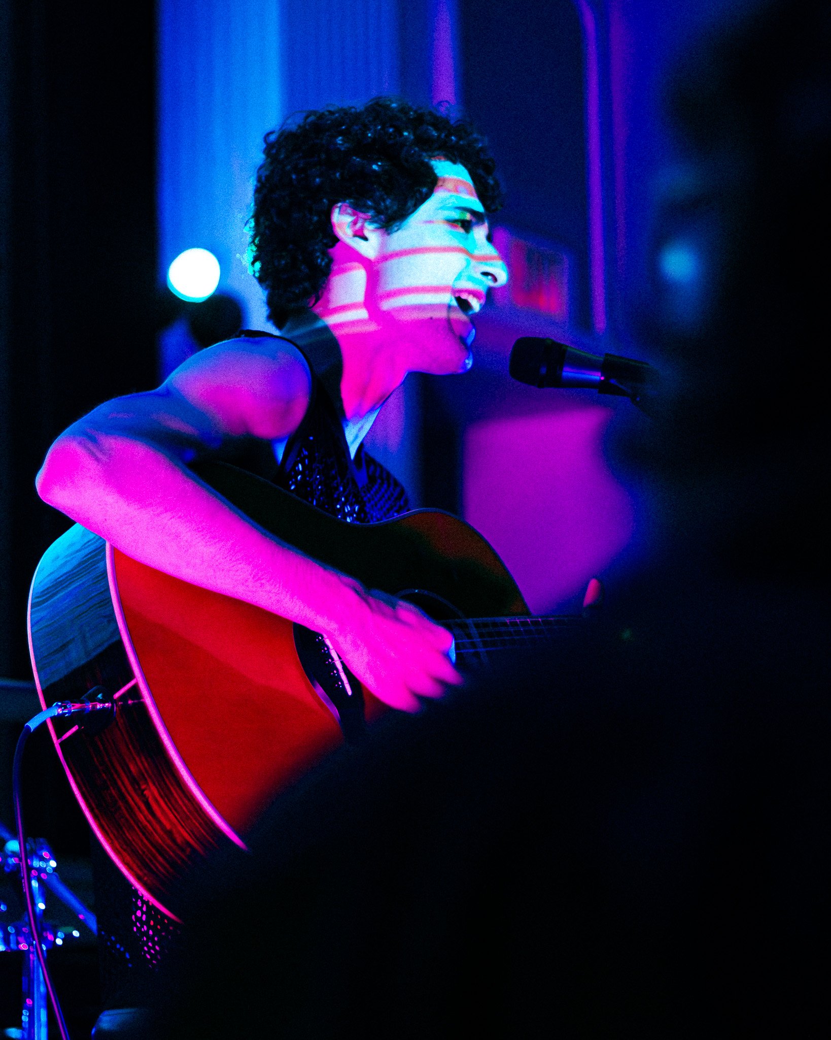 A person with short curly hair singing into a microphone while playing an acoustic guitar, illuminated by colorful blue and pink stage lights.