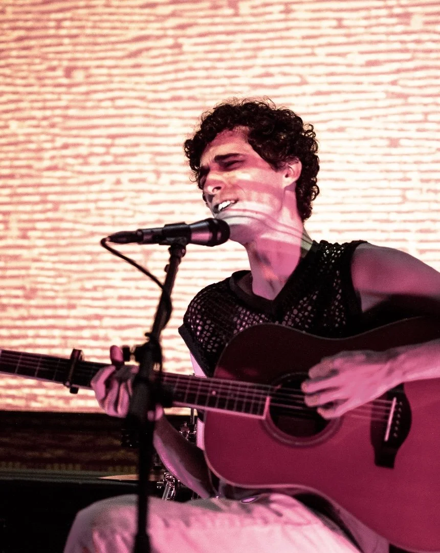 A person with curly hair singing into a microphone while playing an acoustic guitar, with a textured brick wall in the background.