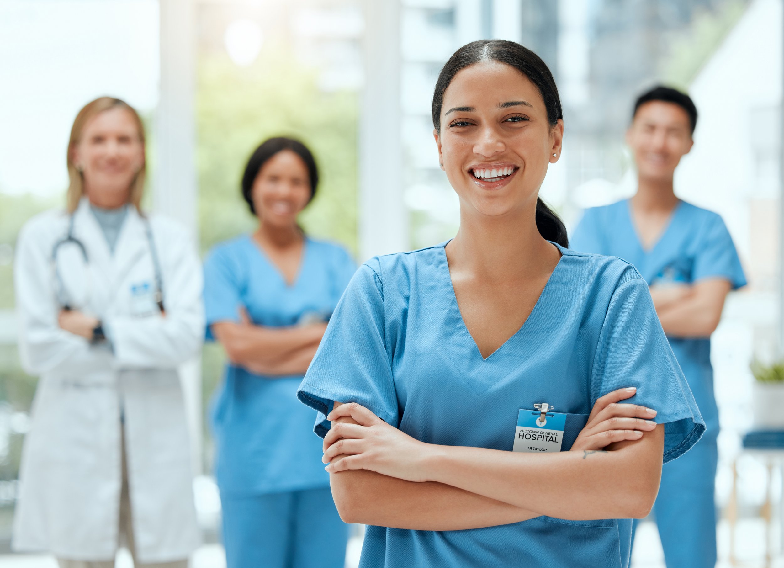 Group of four diverse healthcare professionals in scrubs standing in a hospital. Woman in the forefront with crossed arms smiles at the camera. Behind her, a woman in a white coat with a stethoscope and three women in blue scrubs are visible, all smiling.