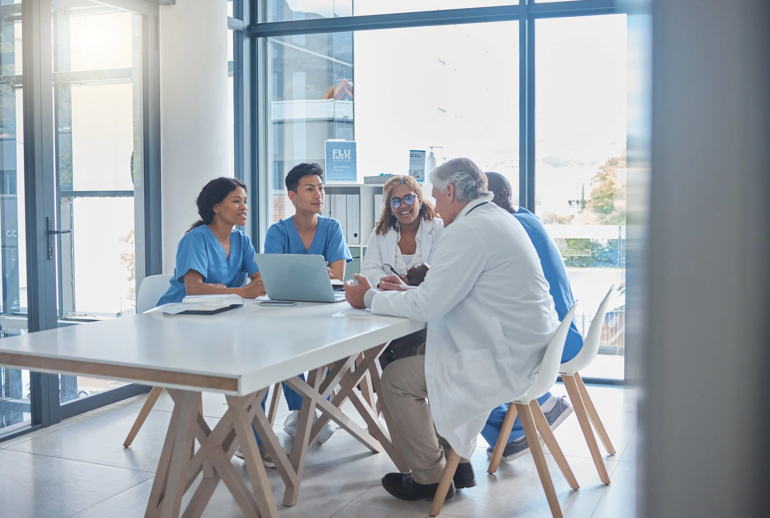 Medical team meeting in a modern clinic. Five healthcare professionals, including doctors and nurses, are seated around a white table in discussion, with large windows and bright natural light in the background.