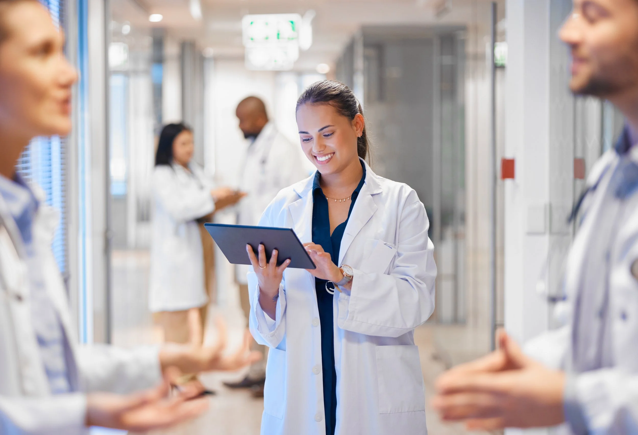 Group of diverse medical professionals in white coats and scrubs in hospital corridor, engaging with digital tablets, smiling and talking.