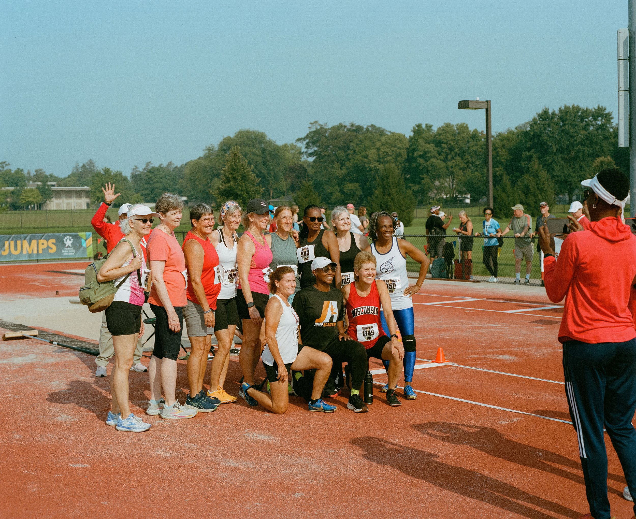 A group of people, mostly women, gathered for a photo on a running track at an outdoor athletic event. They are smiling, some wearing medals, and are dressed in athletic attire. A woman taking a photo is visible on the right side of the image.