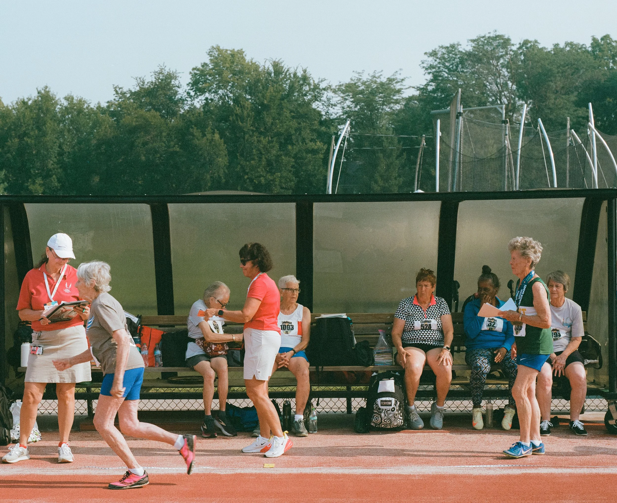 Group of women sitting on a bench and walking on a running track, with some women wearing race bibs, at an outdoor sports facility with trees in the background.