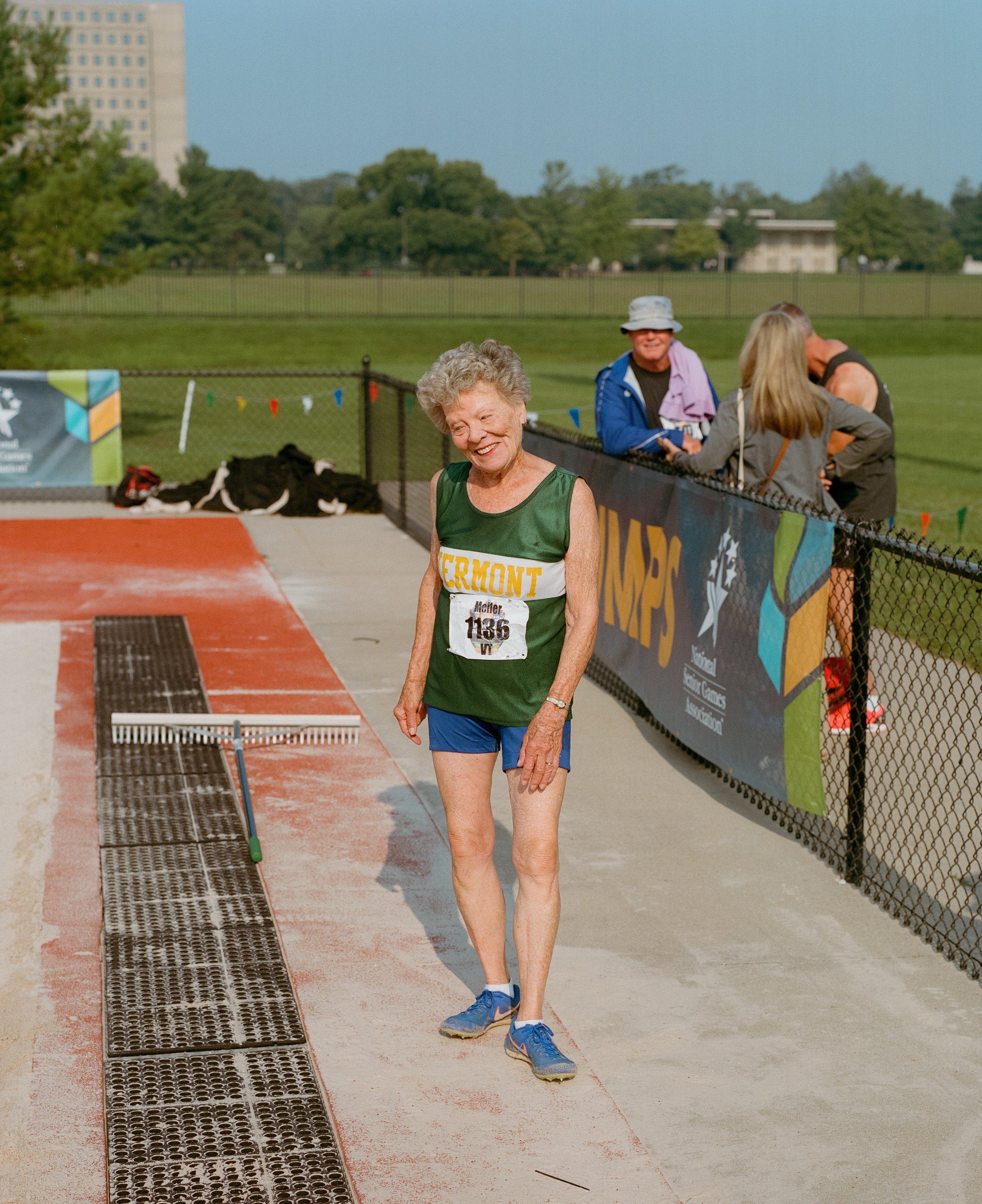 An elderly woman with gray curly hair smiling after a race at a track and field event, wearing a green and yellow Vermont jersey and blue shorts, with a race number 1136 pinned to her shirt, standing on a red track surface near a metal rake.