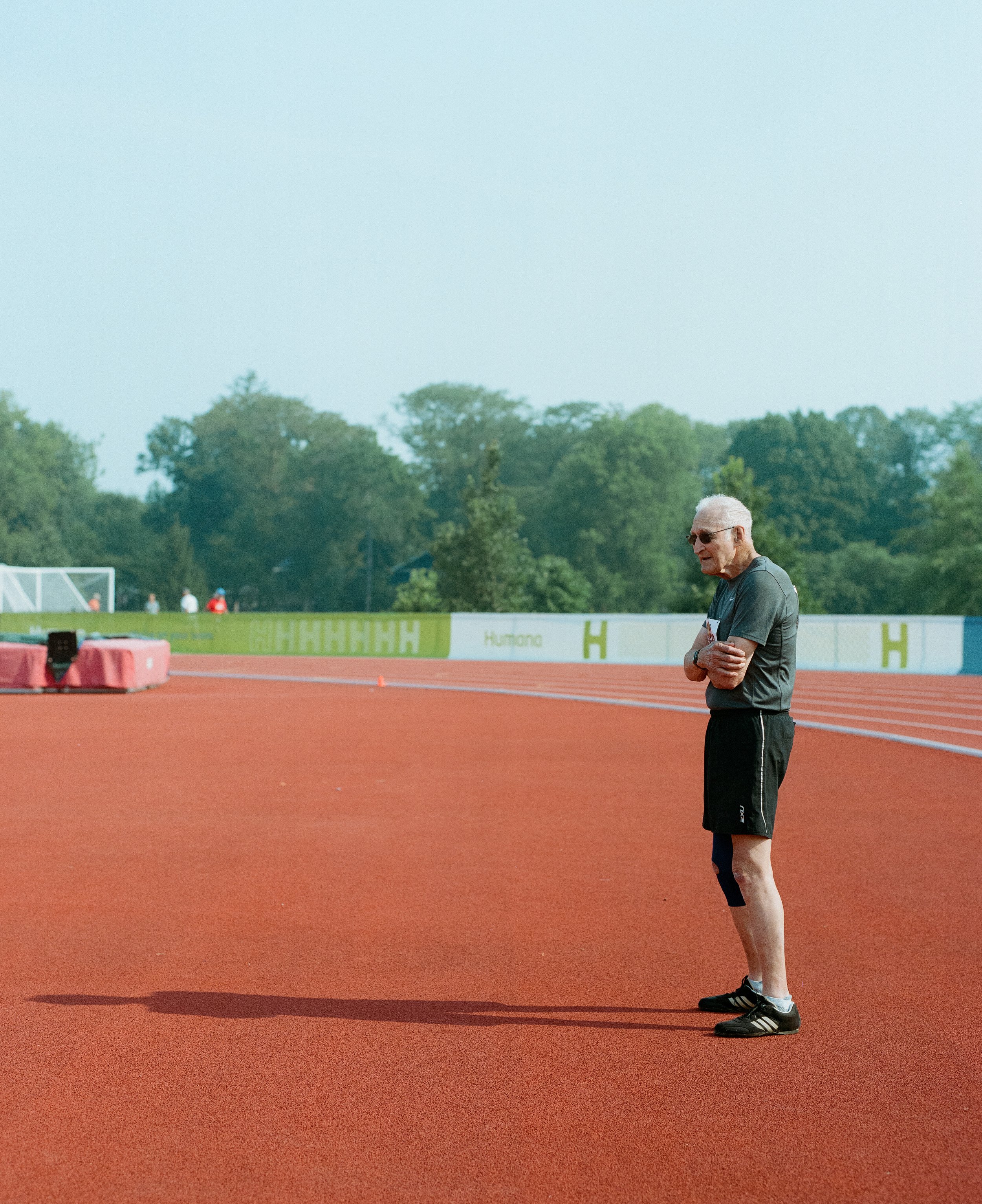 An older man with white hair and sunglasses standing on a red running track at an outdoor sports facility, with trees in the background.