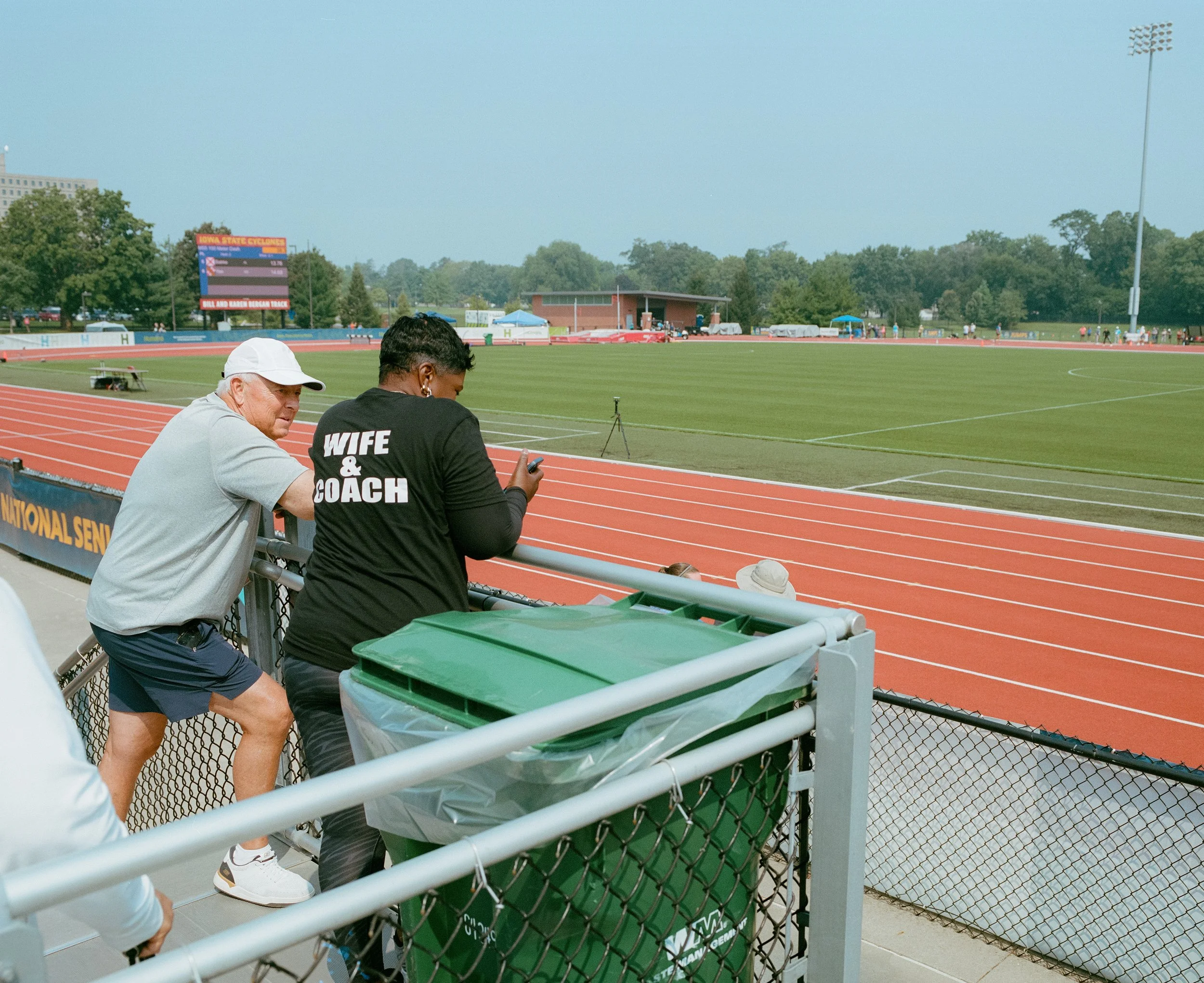 Two people standing by a running track at a stadium, with a green recycling bin in the foreground. One person wears a gray T-shirt and shorts, and the other wears a black shirt with the words 'WIFE & COACH'. The track surrounds a green field, with a 