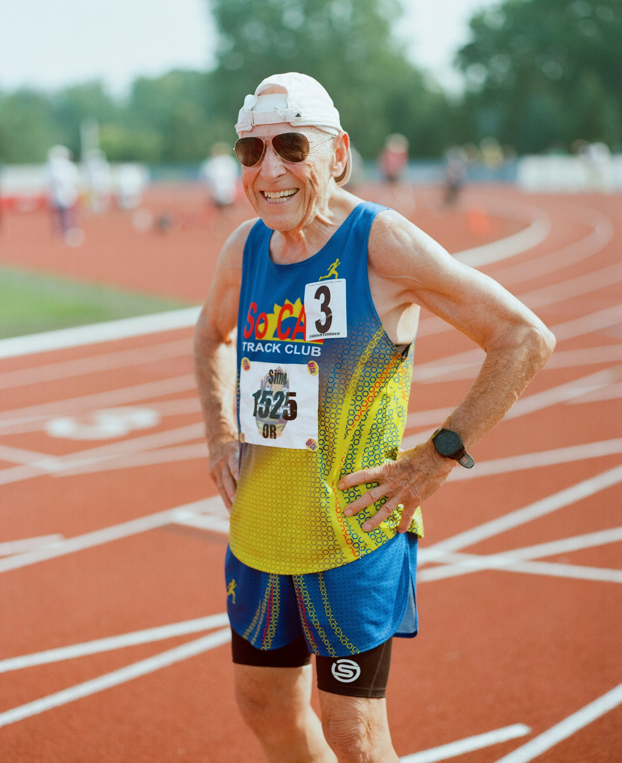 Elderly male runner smiling on track wearing athletic gear, sunglasses, and a white cap, with race bib number 1525, during a track event.