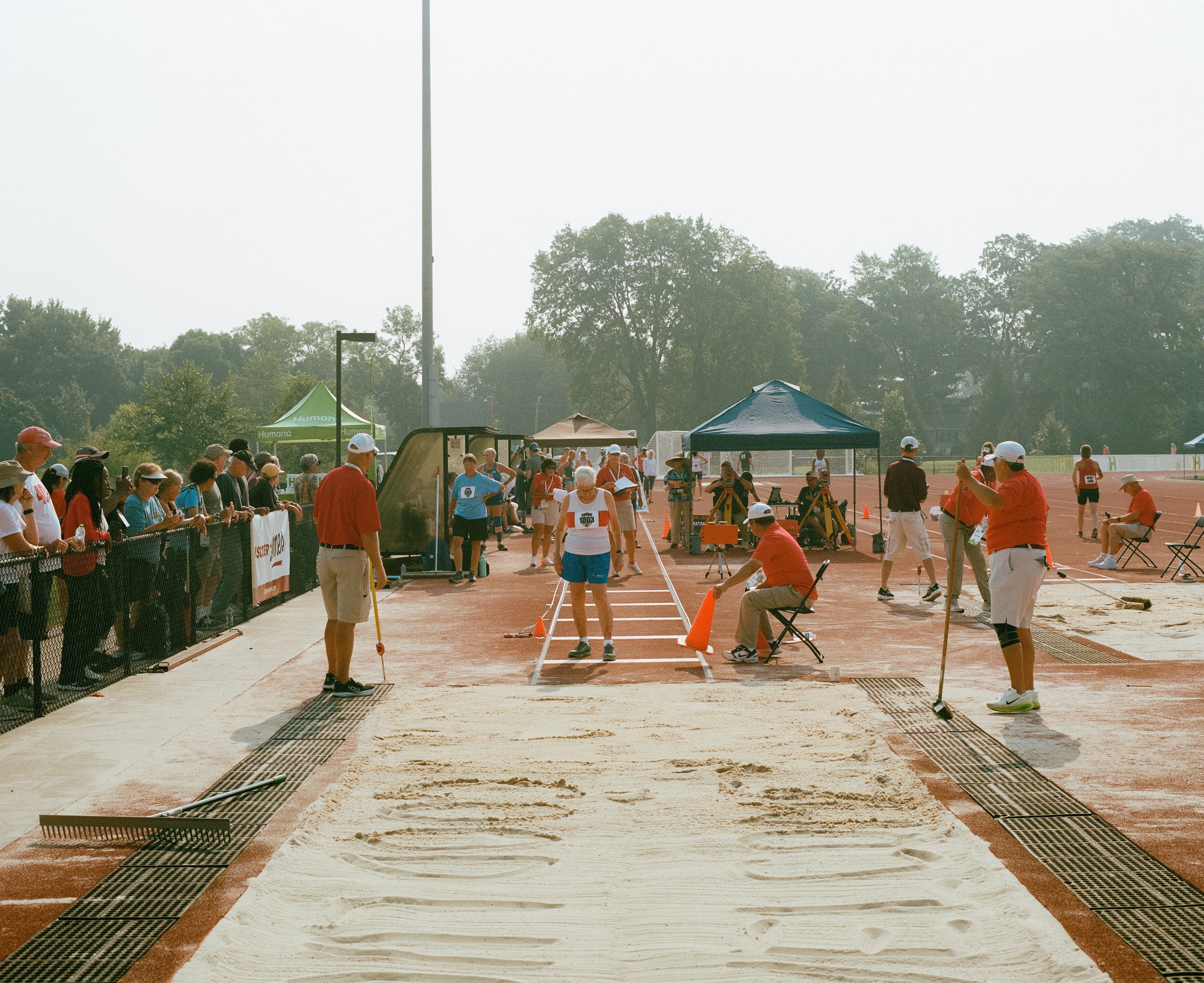 Long jump pit with athletes and officials preparing for a jump at a track and field event, surrounded by spectators and tents.