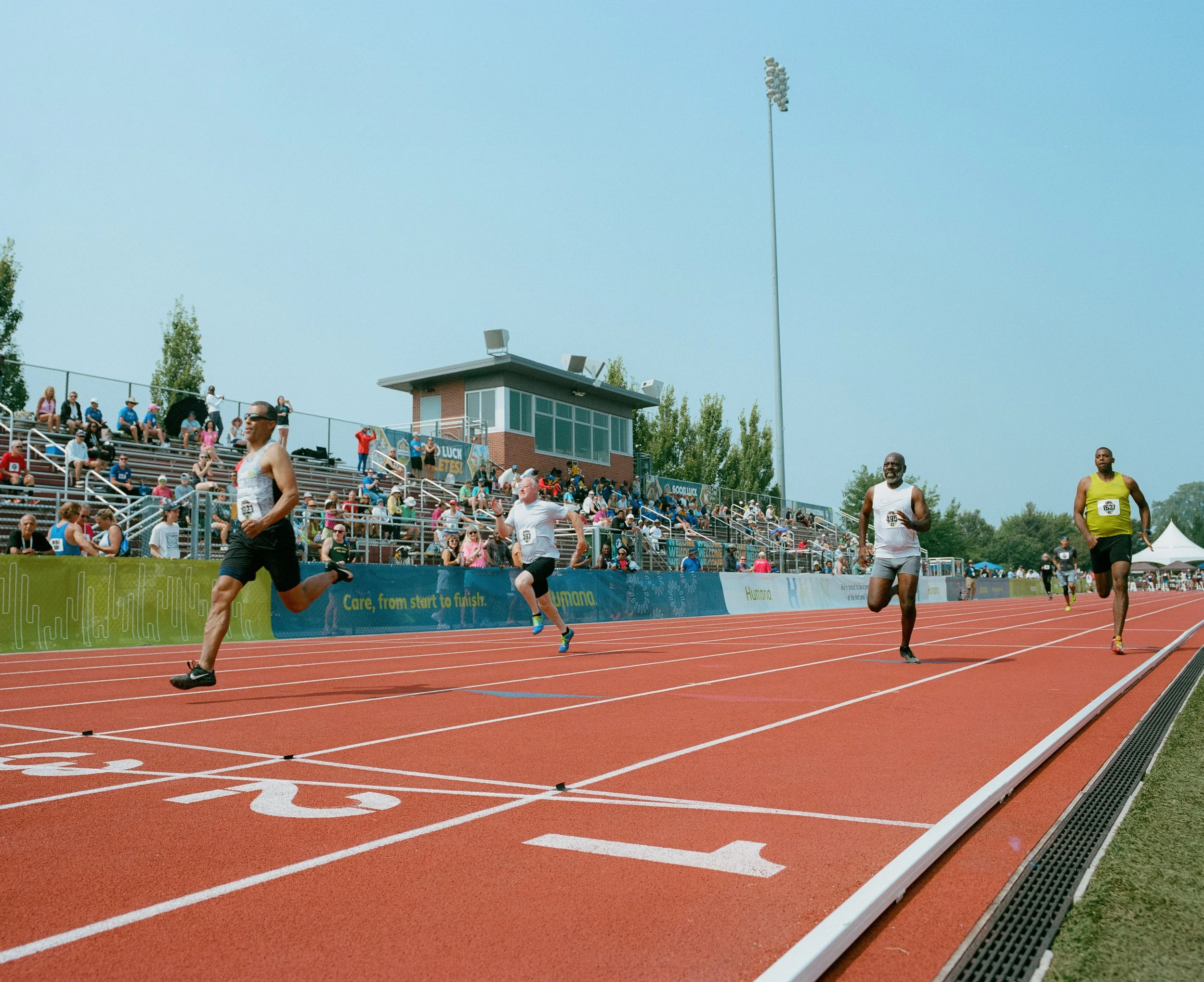 A group of male runners competing in a race on an outdoor track with spectators sitting on bleachers in the background.
