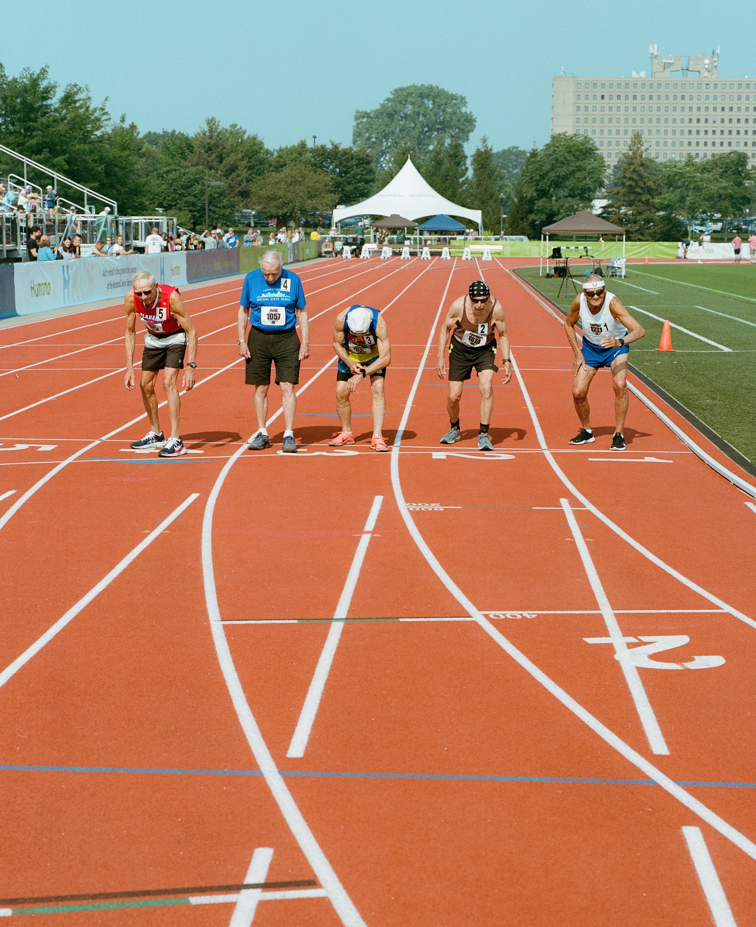 Track athletes starting a race at the starting line on a bright, sunny day, with a crowd and trees in the background.