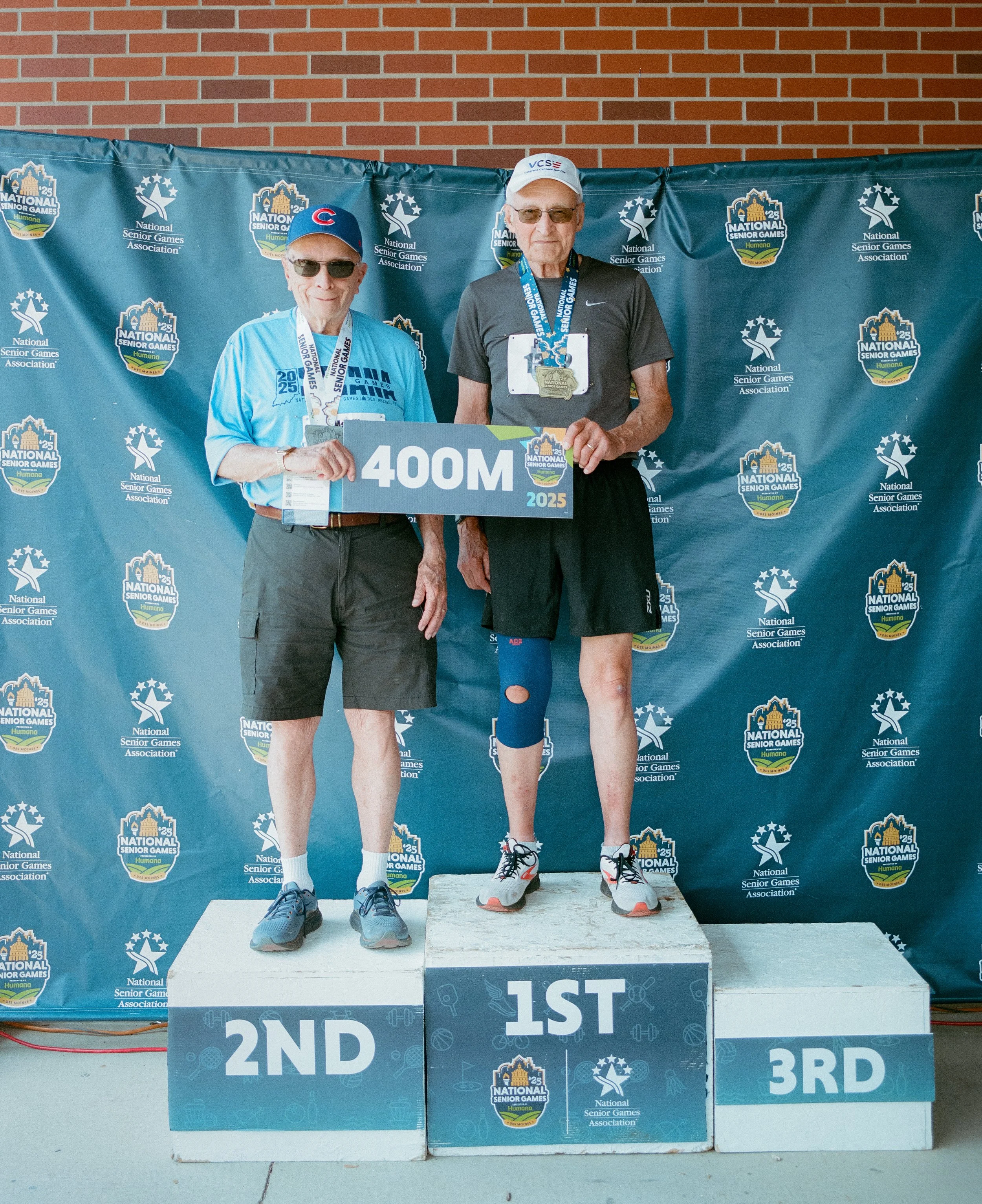 Two elderly men standing on a winners' podium at a senior games event, with medals around their necks. One man is holding a sign that reads '400M 2025'. The background features posters and logos for the National Senior Games Association.
