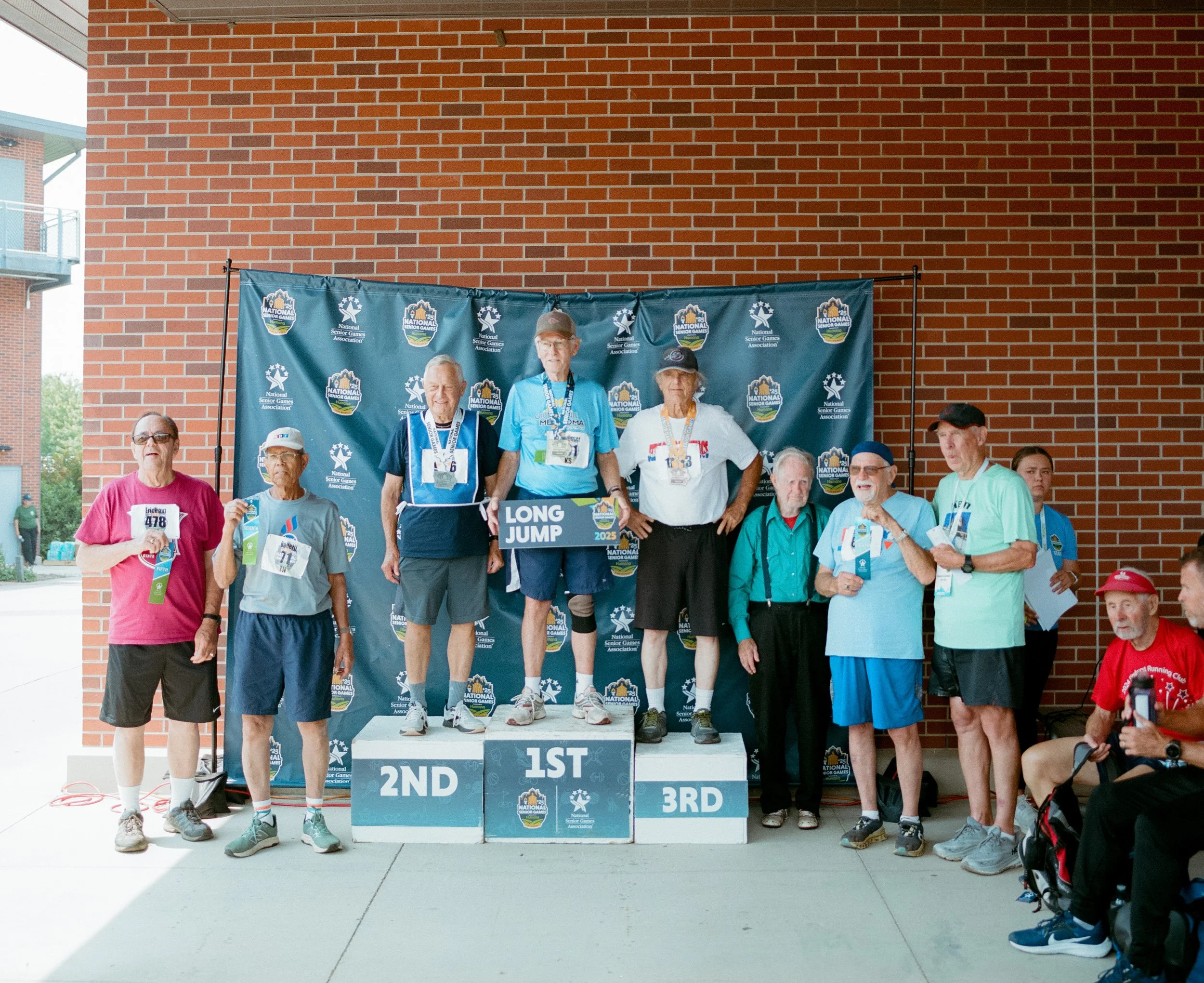 Group of elderly men and women on a winners' podium at a race event, with three top finishers standing on the 1st, 2nd, and 3rd place platforms, holding medals and a sign that says 'LONG JUMP 2023.' The background features a blue banner with event lo