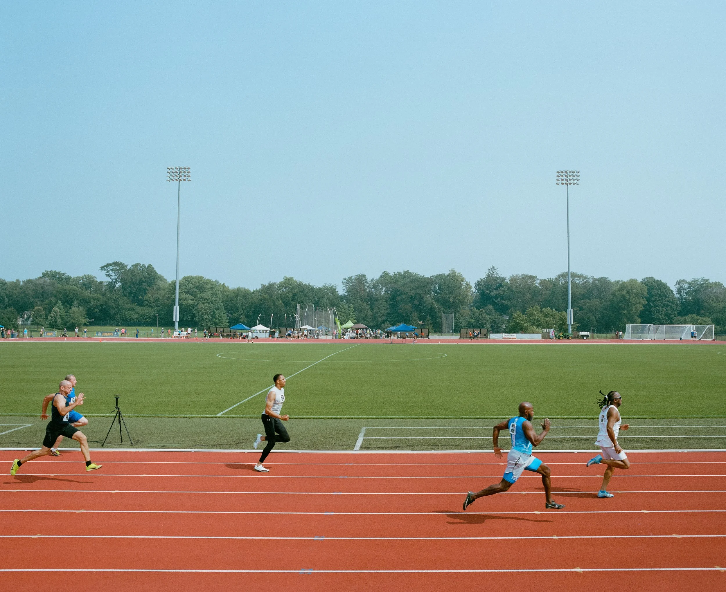 Male athletes running on a red track during a race at an outdoor stadium with a green field, trees, and tall floodlights in the background.