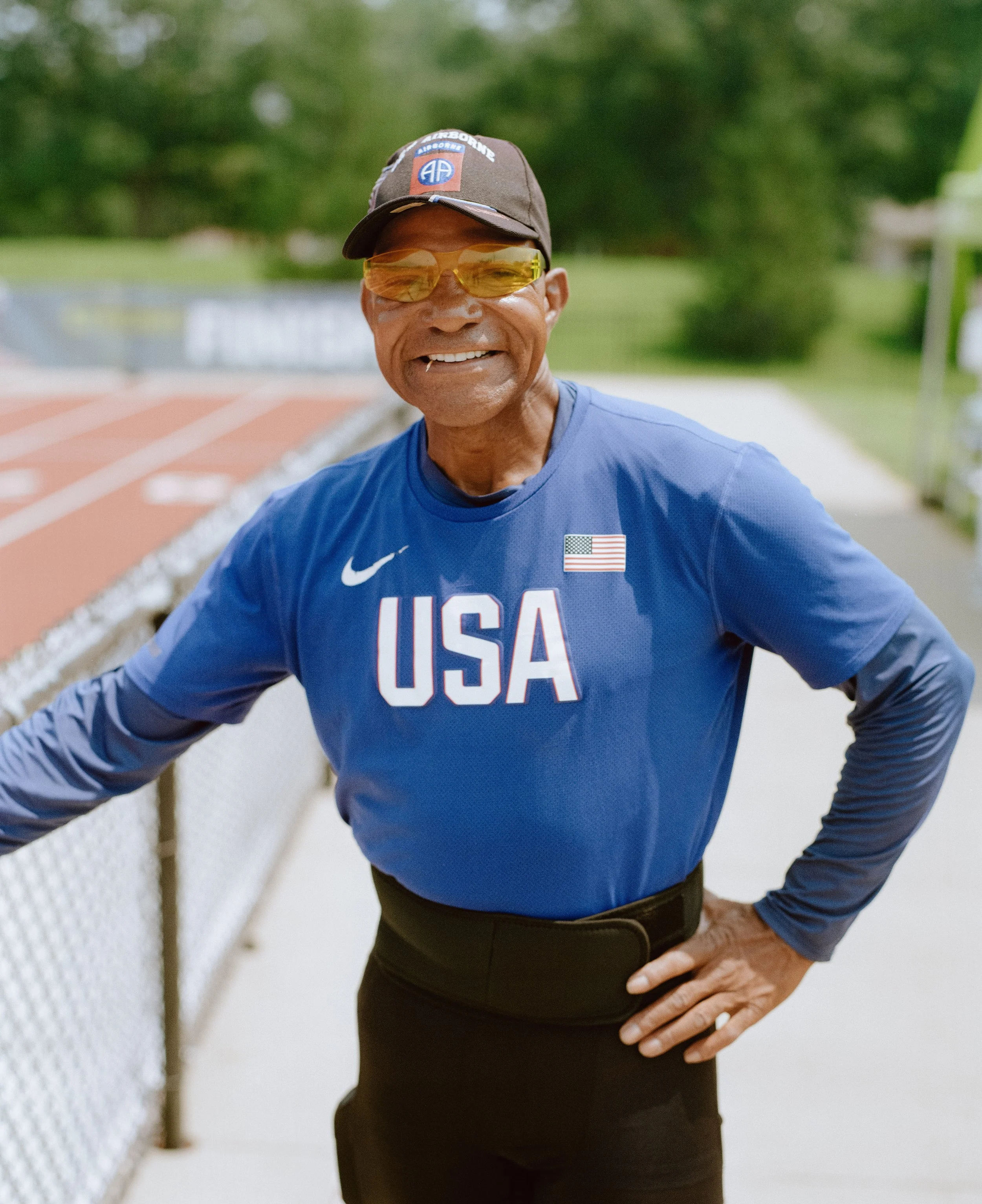 An elderly man standing outdoors by a fence, smiling, wearing a blue USA athletic shirt, a cap, and sunglasses with a track and green trees in the background.