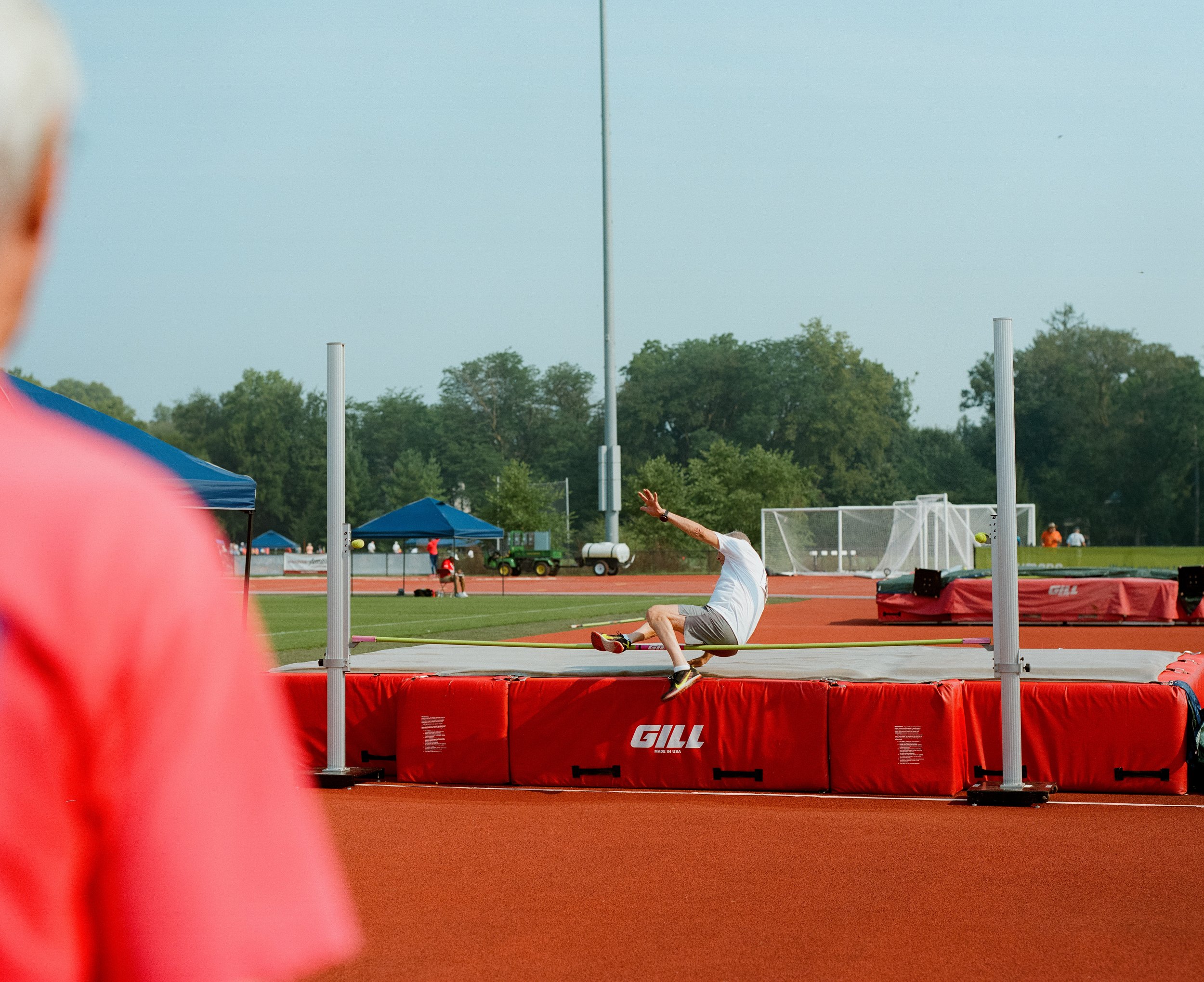 An athlete in white shirt and gray shorts attempting the long jump at an outdoor track and field event. The scene shows a red cushioned landing area, a running track, and a background of trees and a blue sky.