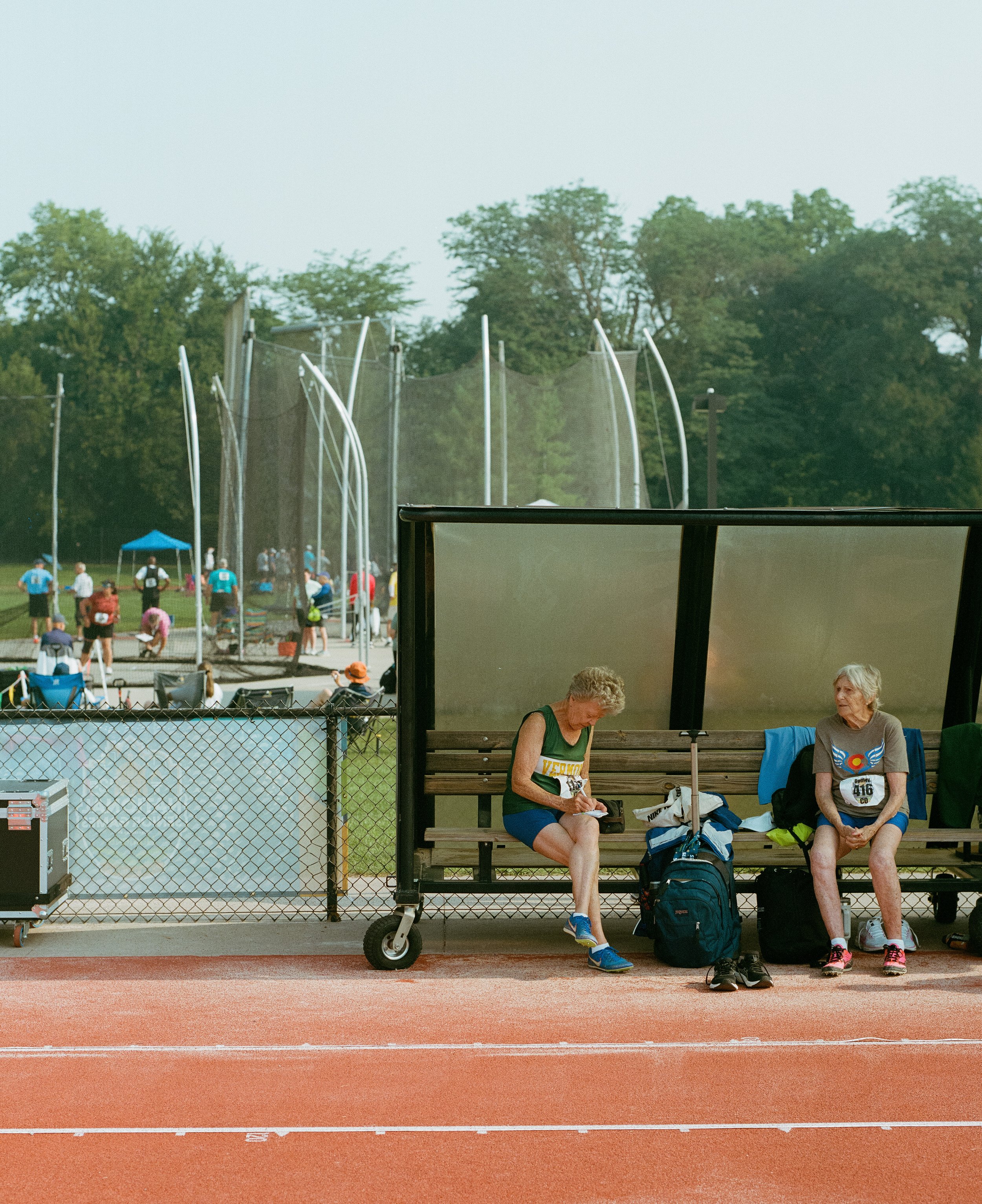Two women sitting on a bench at a track and field stadium, waiting during a race, with other athletes and officials in the background near the throwing area.