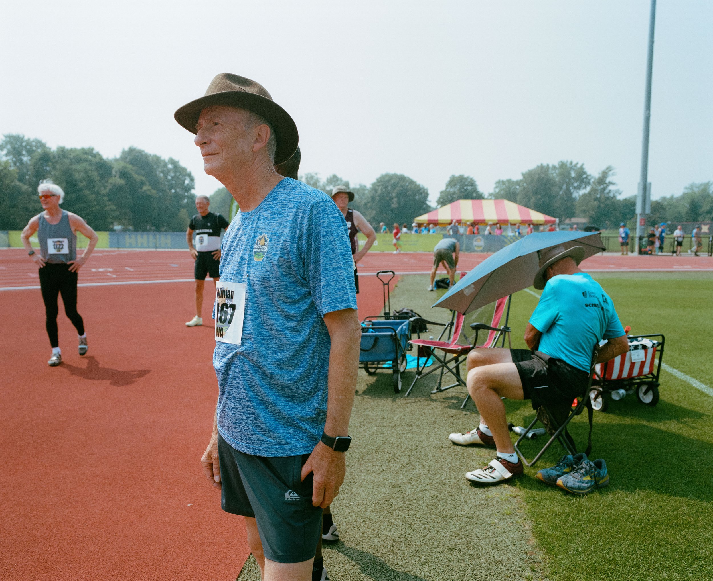 An elderly man in athletic clothing stands on a track at a sporting event, wearing a gray hat and a blue shirt with a race number. Other athletes and officials are in the background, with some sitting under umbrellas on the grassy area beside the tra