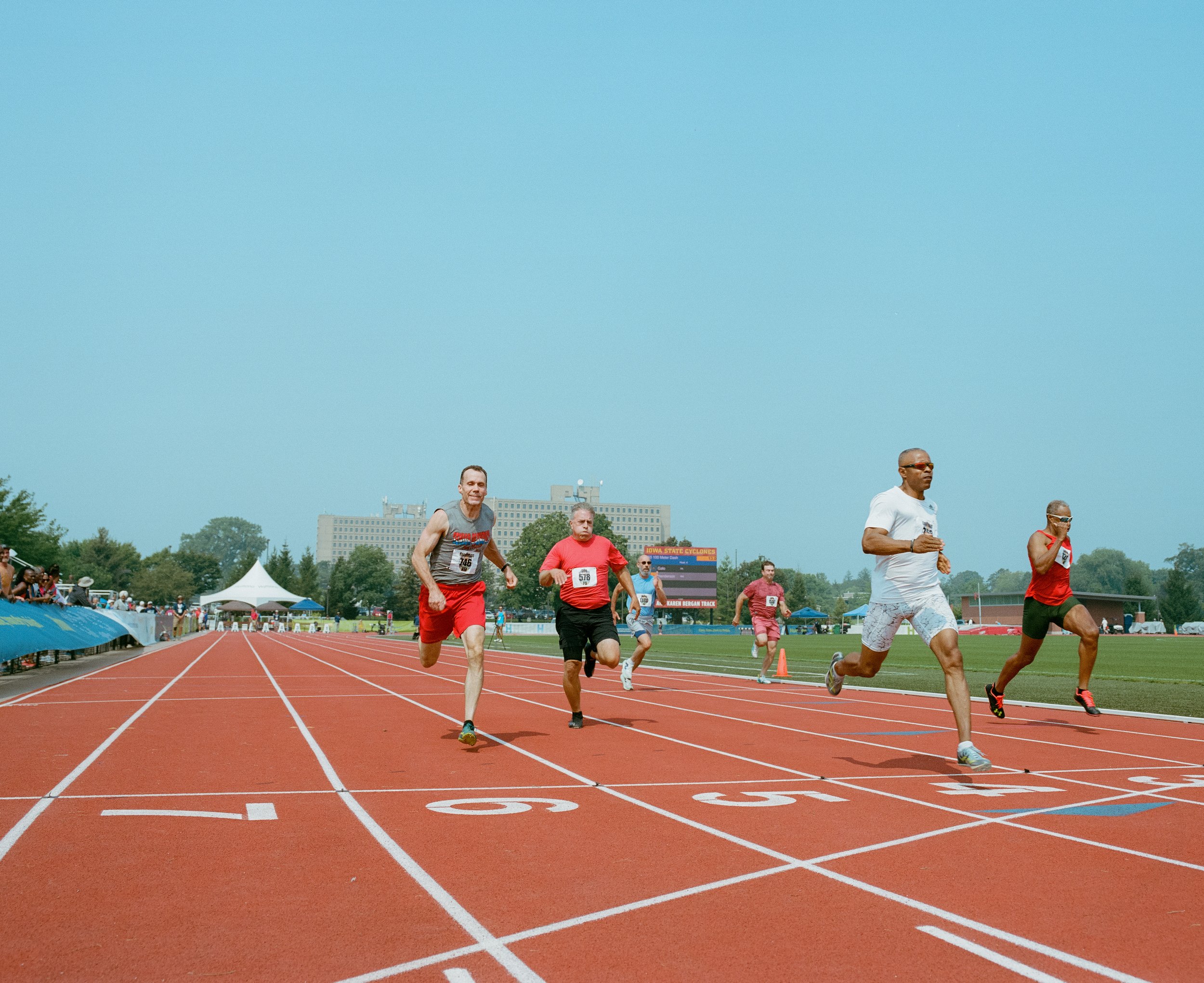 People running on a track during a race at an outdoor stadium on a sunny day.