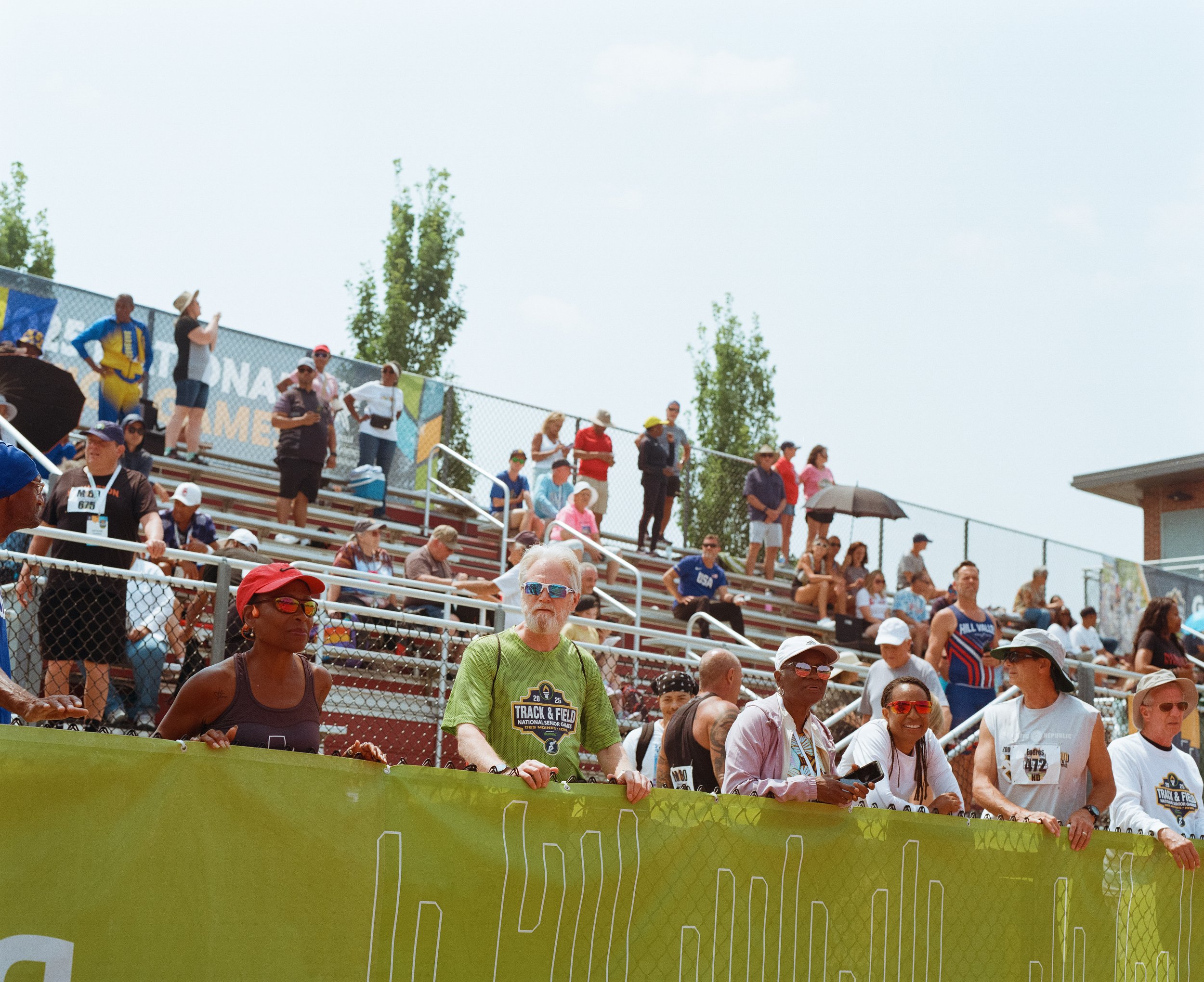 Crowd of spectators at an outdoor athletic event, some sitting on bleachers, others standing, holding umbrellas and wearing sports attire.