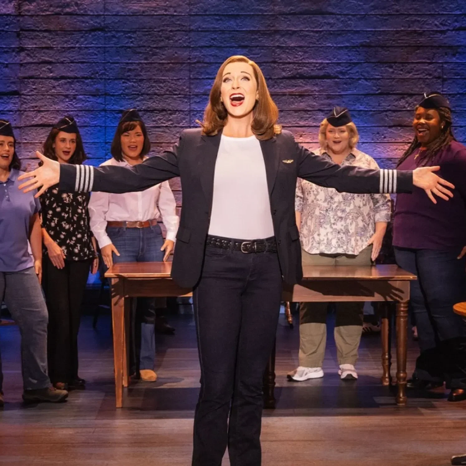 A woman singing with arms outstretched on stage, surrounded by people wearing flight attendant caps.