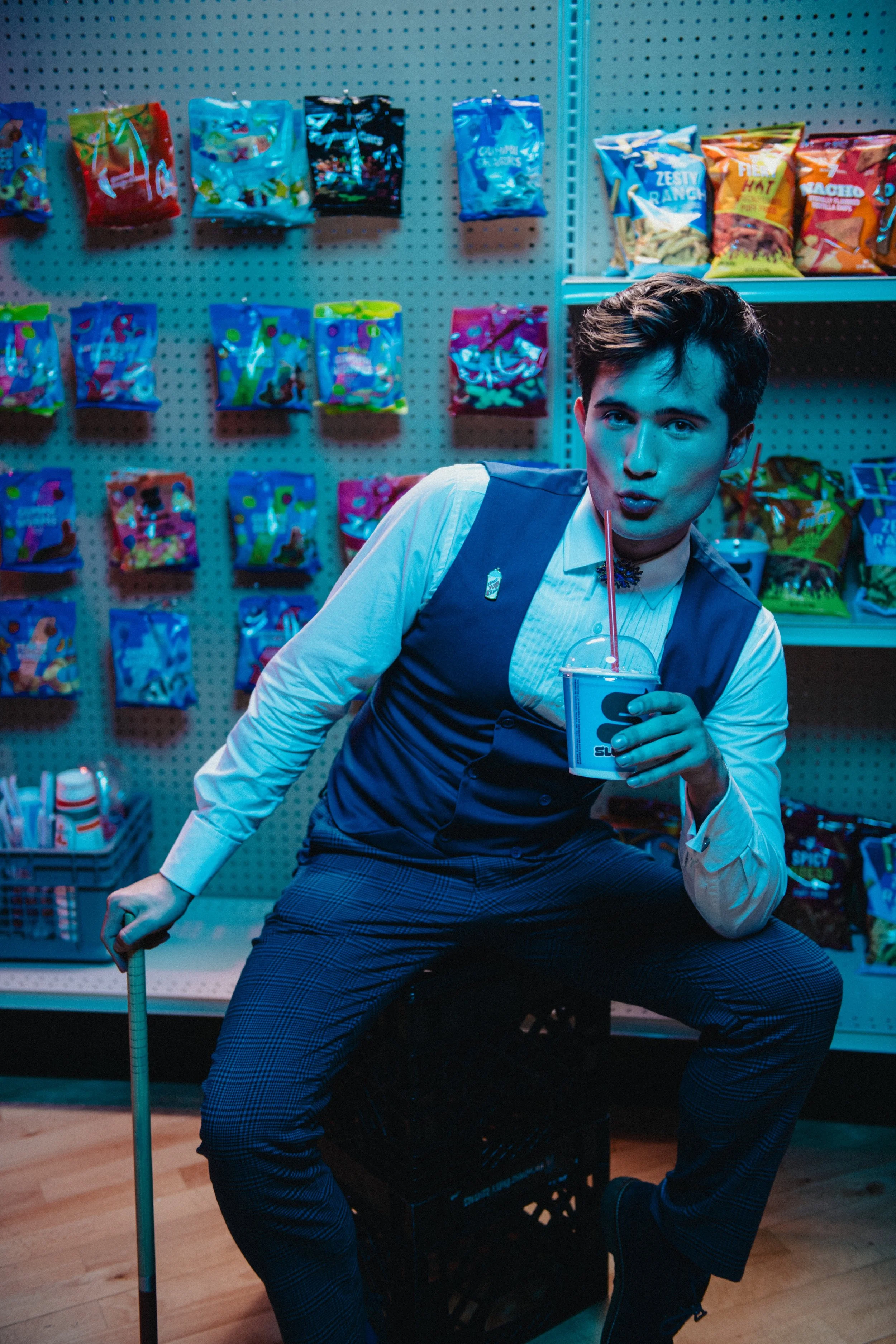 Associate Producer Mickey White at the opening night of Heathers Off-Broadway - sitting on a milk crate in a store aisle with snack bags on the shelves behind them, holding a drink with a straw and making a playful expression.