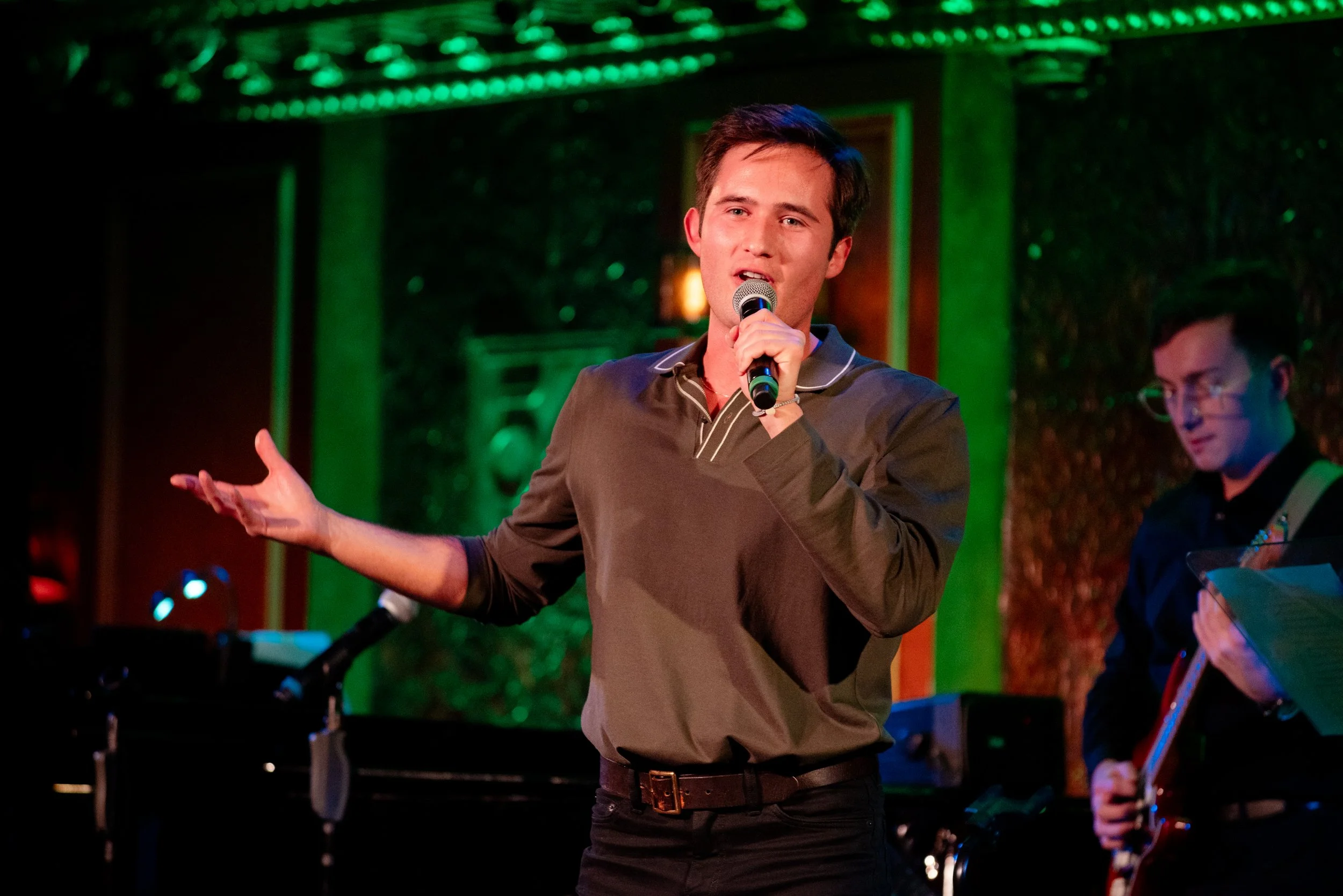 Mickey White Performing at 54 Below with a musician playing guitar in the background, and colorful stage lighting in NYC.