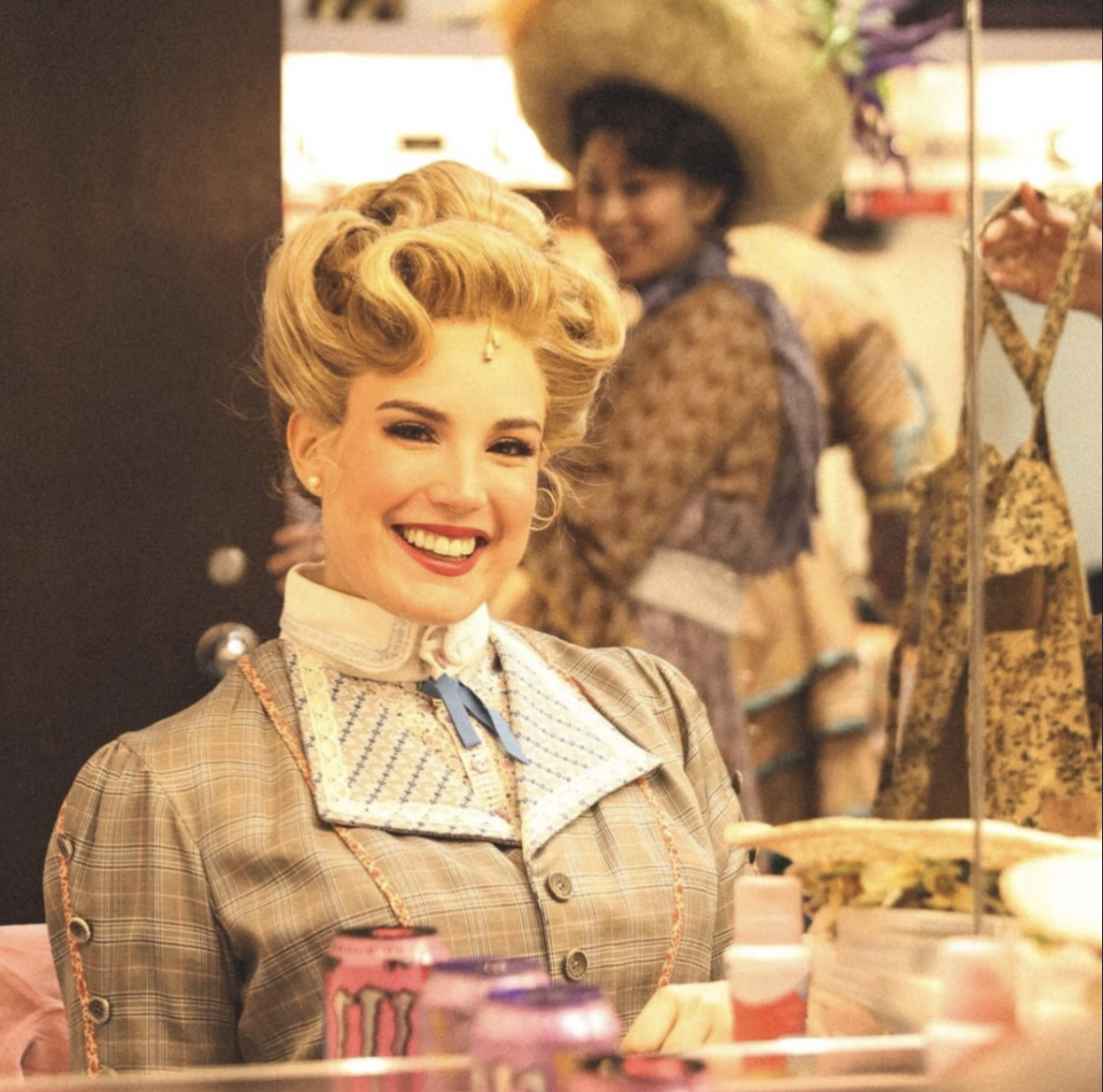 A woman dressed in vintage clothing with her hair styled in curls, smiling at a vanity mirror. In the background, another woman wearing a large hat and similar vintage attire is visible.