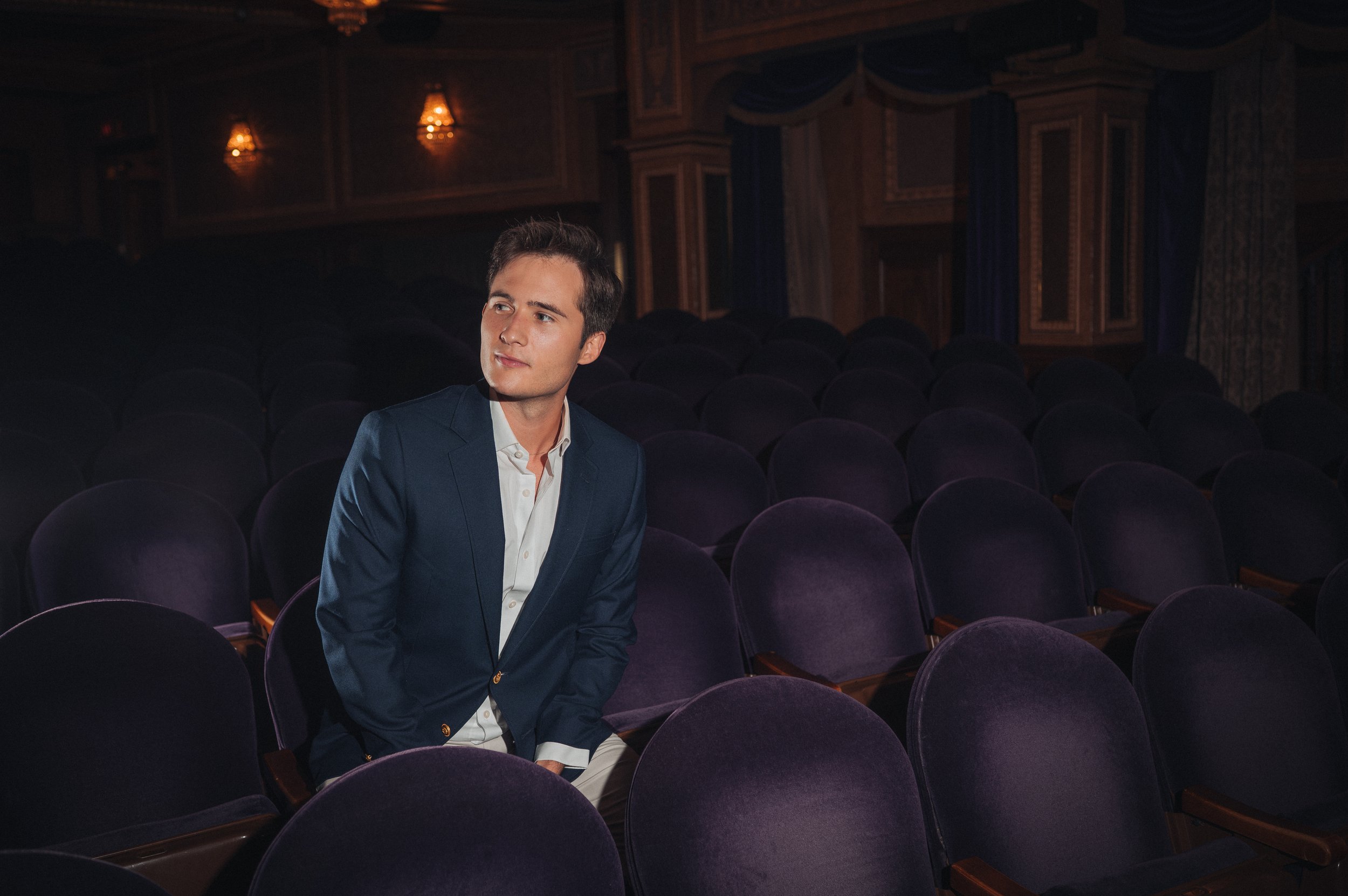 Broadway Producer Mickey White in a blue blazer and white shirt sitting alone in an empty theater with purple seats and warm lighting.