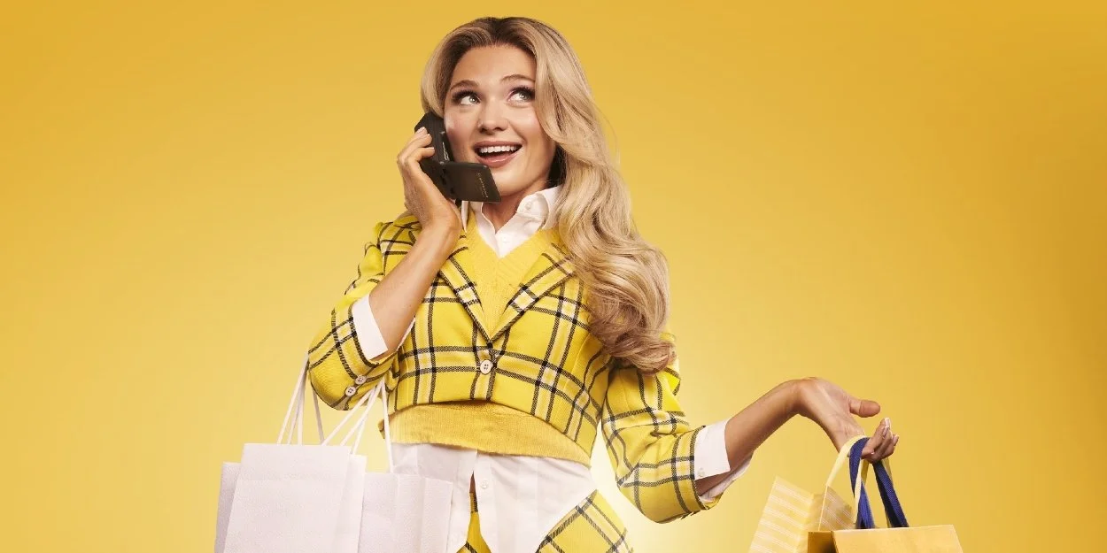 A young woman with long blonde hair, wearing a yellow plaid blazer and white shirt, talking on a cell phone while holding shopping bags against a solid yellow background.