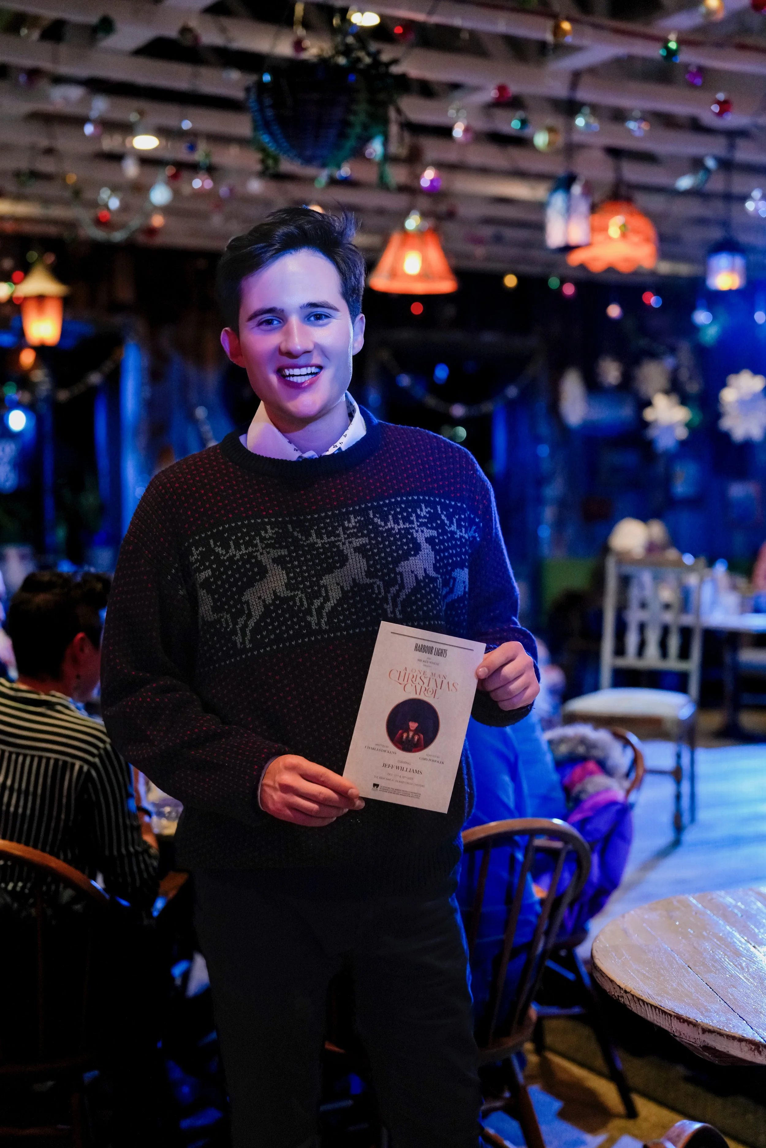 A young man smiling at a Christmas gathering, holding a program or flyer, in a decorated, cozy indoor setting with festive decorations and other people seated at tables in the background.