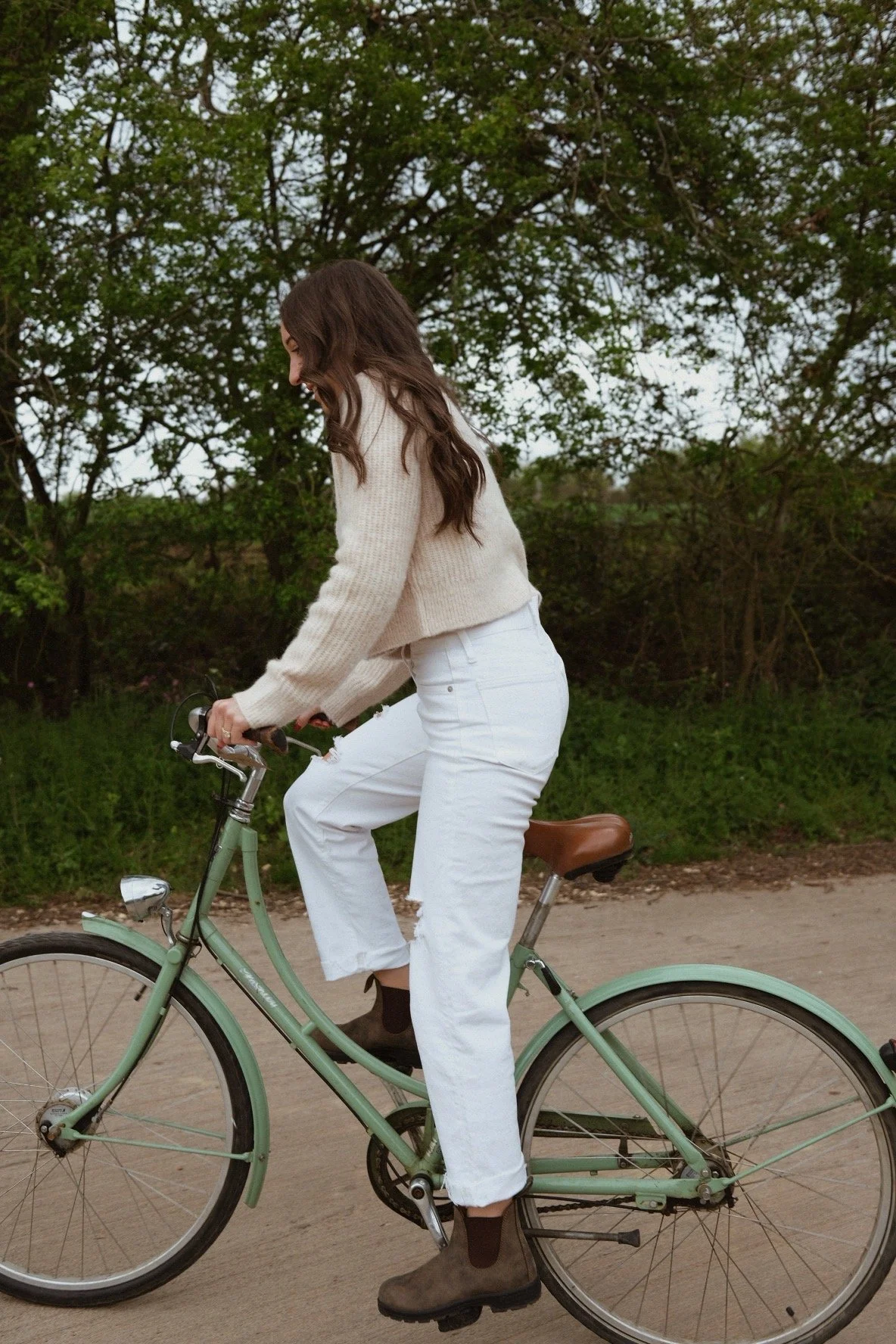 A woman with long brown hair riding a green bicycle on a dirt path surrounded by greenery and trees.
