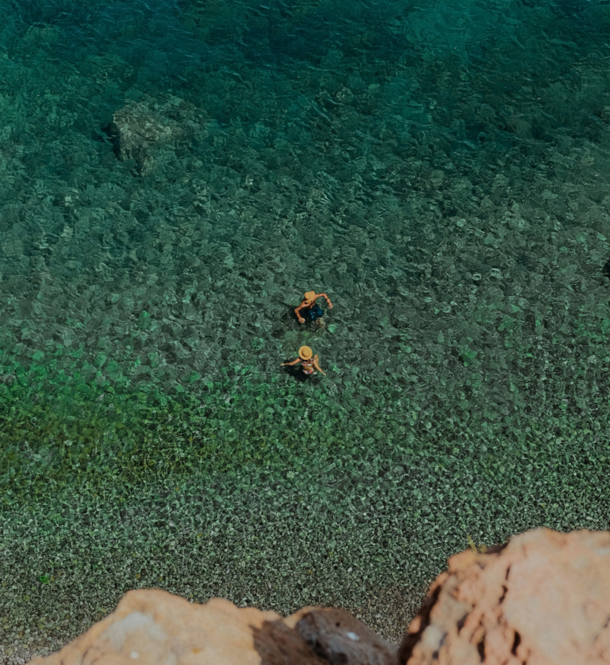 Two people swimming in a clear, green body of water near the shoreline seen from above.